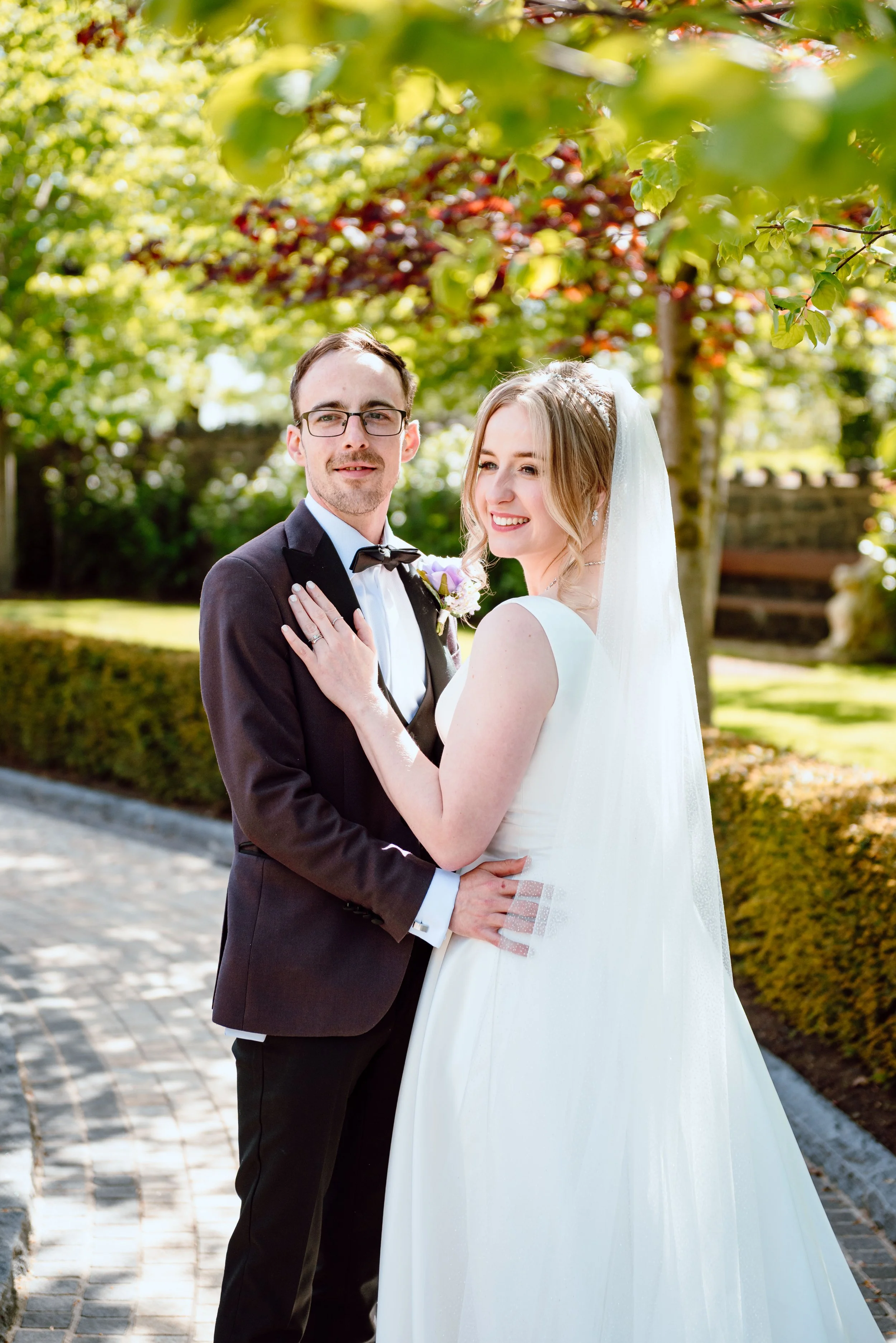 A bride and groom posing outdoors on their wedding day, with trees and a garden in the background. The bride is wearing a white wedding gown with a veil, and the groom is dressed in a black tuxedo with a bow tie.
