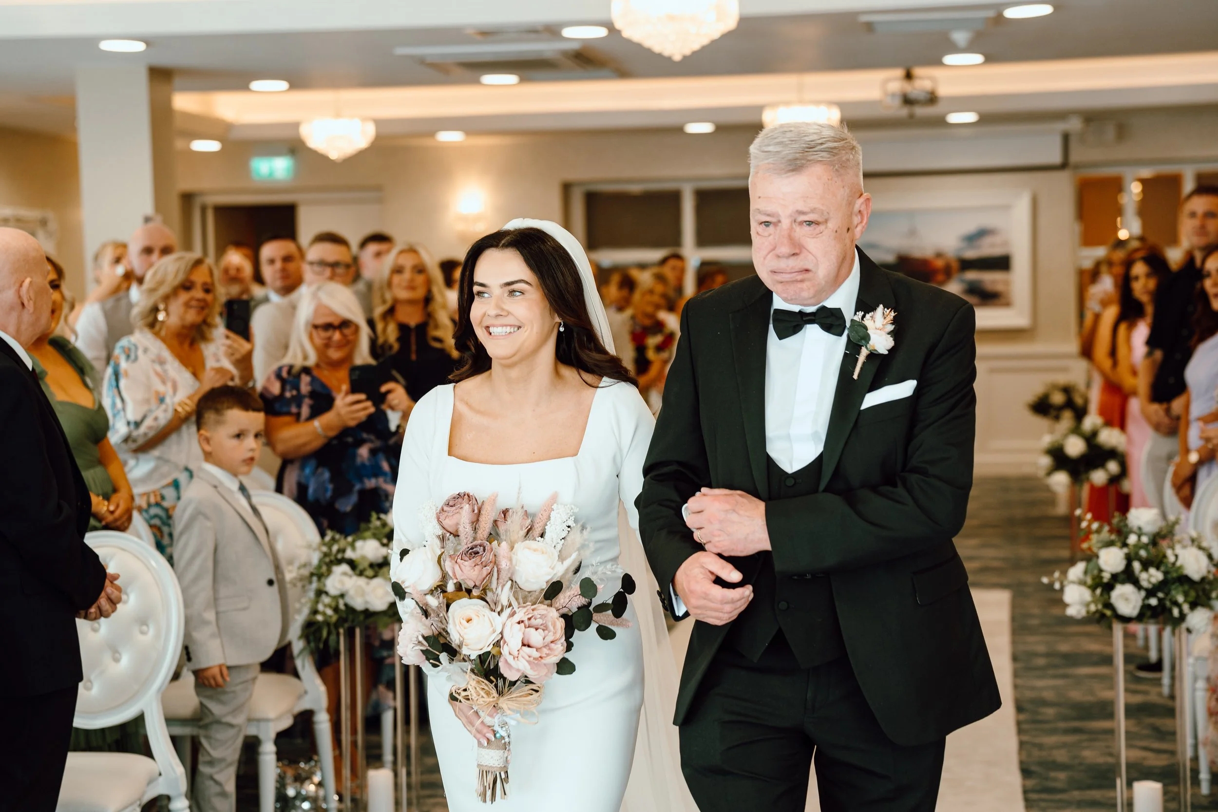 A bride in a white dress holding a bouquet of pink and white flowers, walking down the aisle with an older man in a black tuxedo, at a wedding ceremony with guests watching.