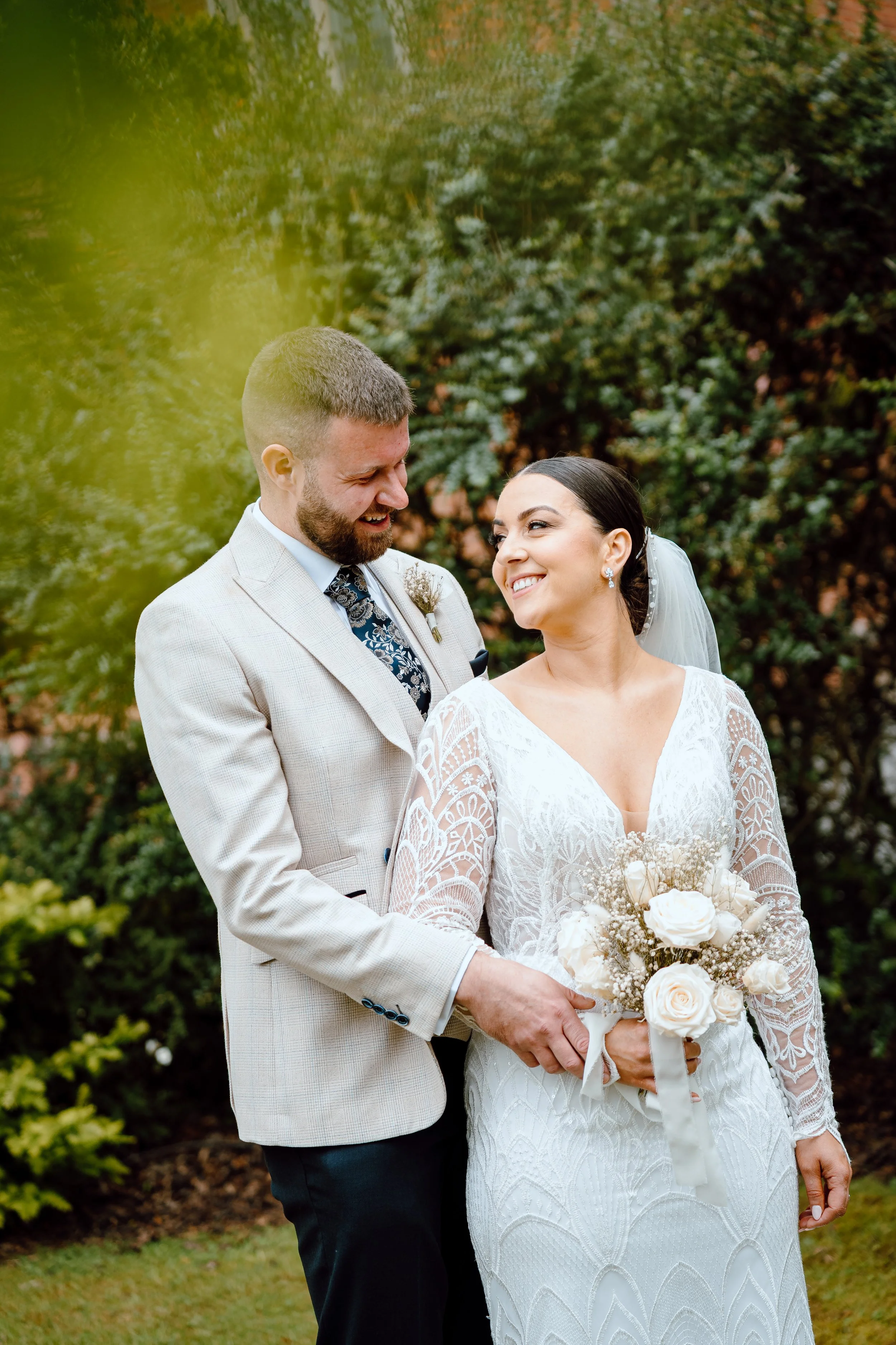 A bride and groom smiling at each other outdoors, the bride holding a bouquet of white roses, surrounded by green foliage.