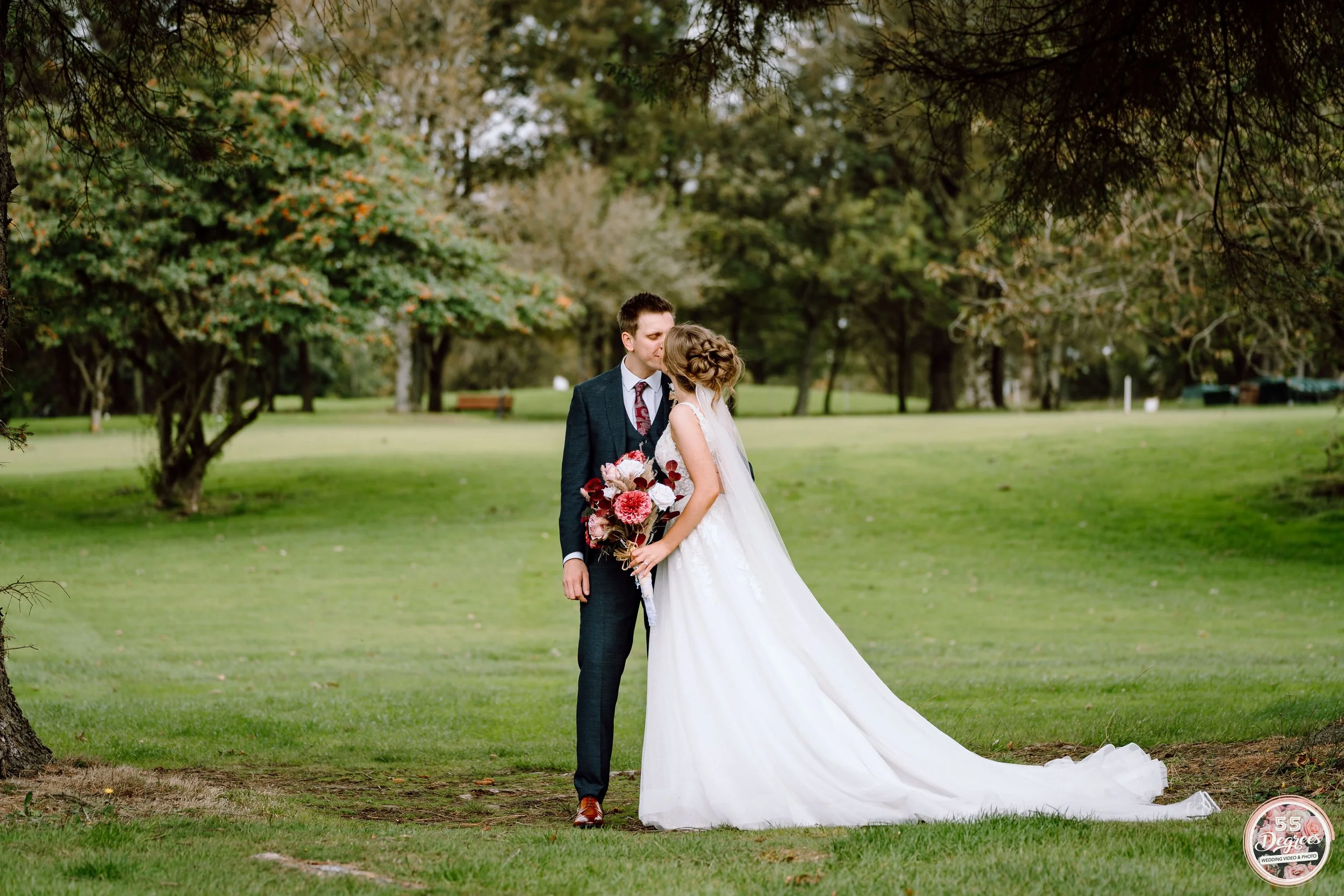 A bride and groom share a kiss in a park, surrounded by green trees and grass. The bride wears a white wedding gown and holds a bouquet, while the groom wears a dark suit.
