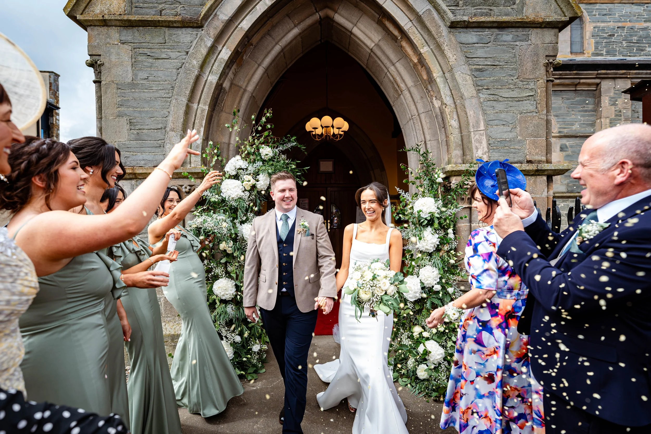 A newly married couple walking out of a church gate, surrounded by smiling friends throwing confetti, as the bride holds a bouquet and the groom wears a tan suit with a boutonniere.