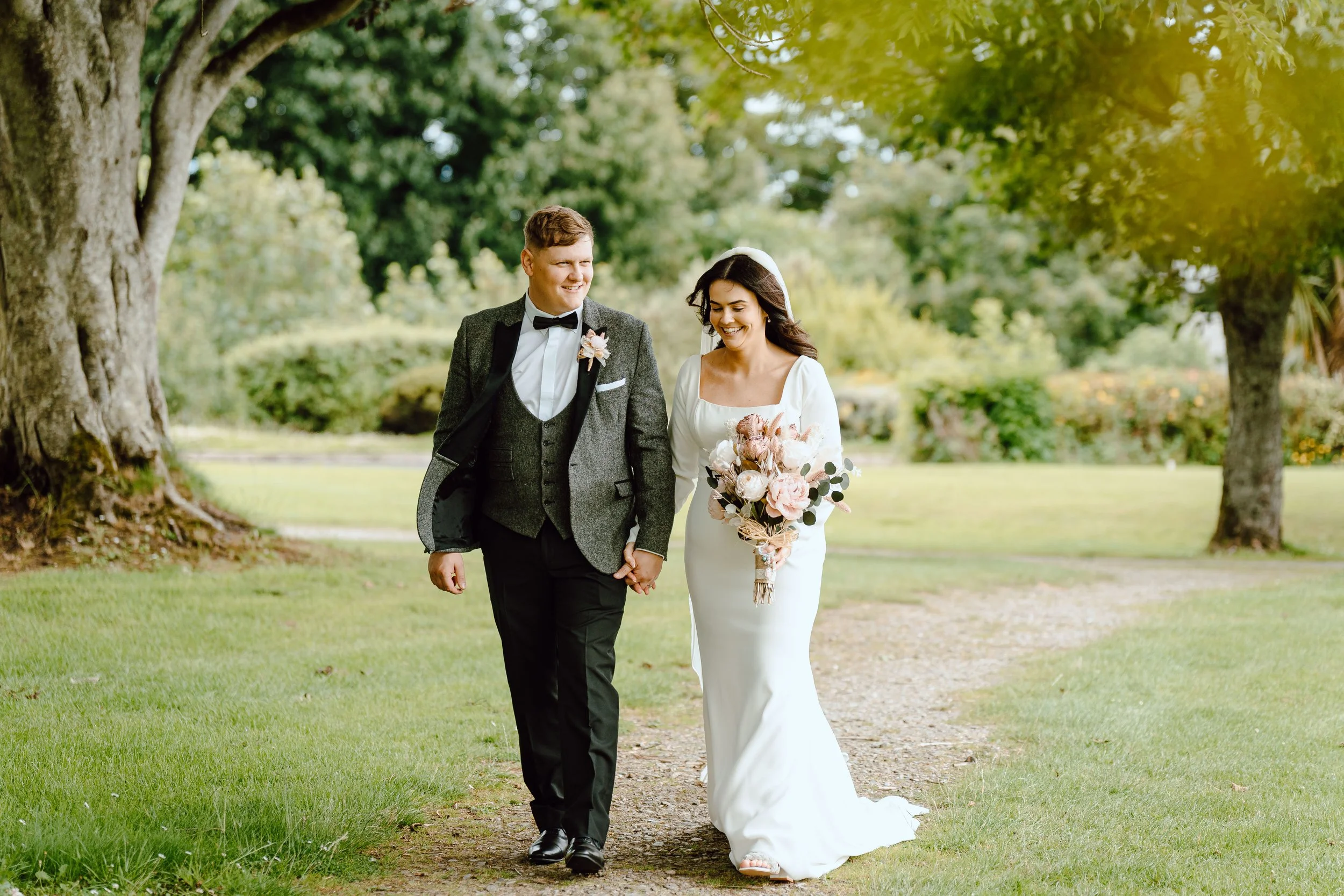 A bride and groom walking hand in hand outdoors on a grassy path surrounded by trees, smiling at each other. The bride is in a white wedding dress holding a bouquet of flowers, and the groom is in a dark tuxedo with a bow tie.