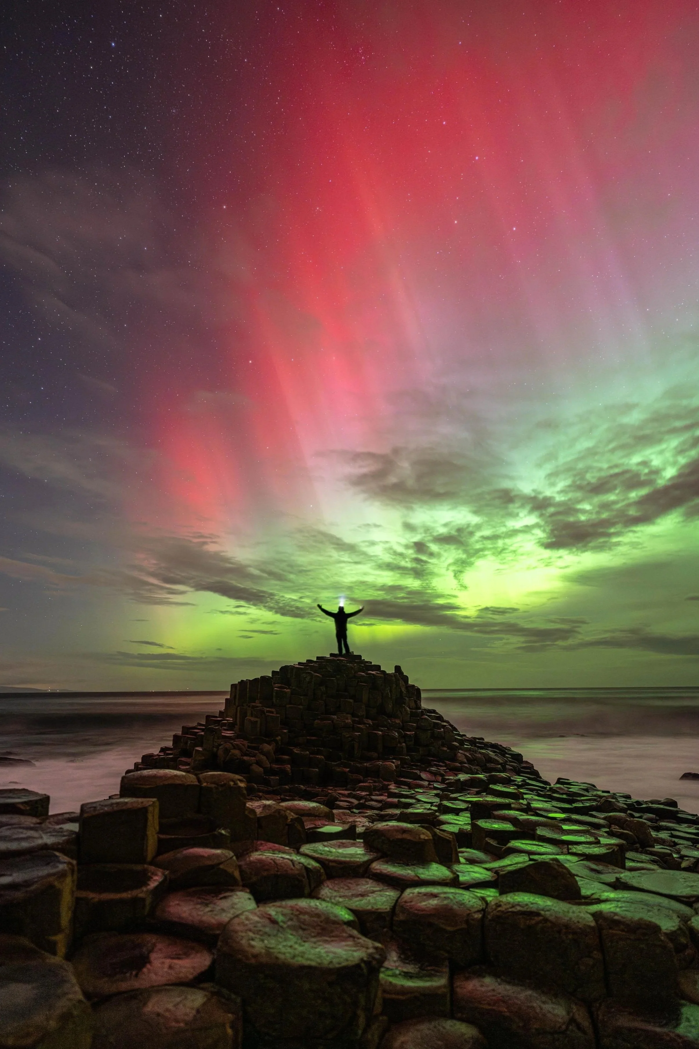 Person standing on a stone pier with arms raised, looking at the Northern Lights in the night sky over the ocean.