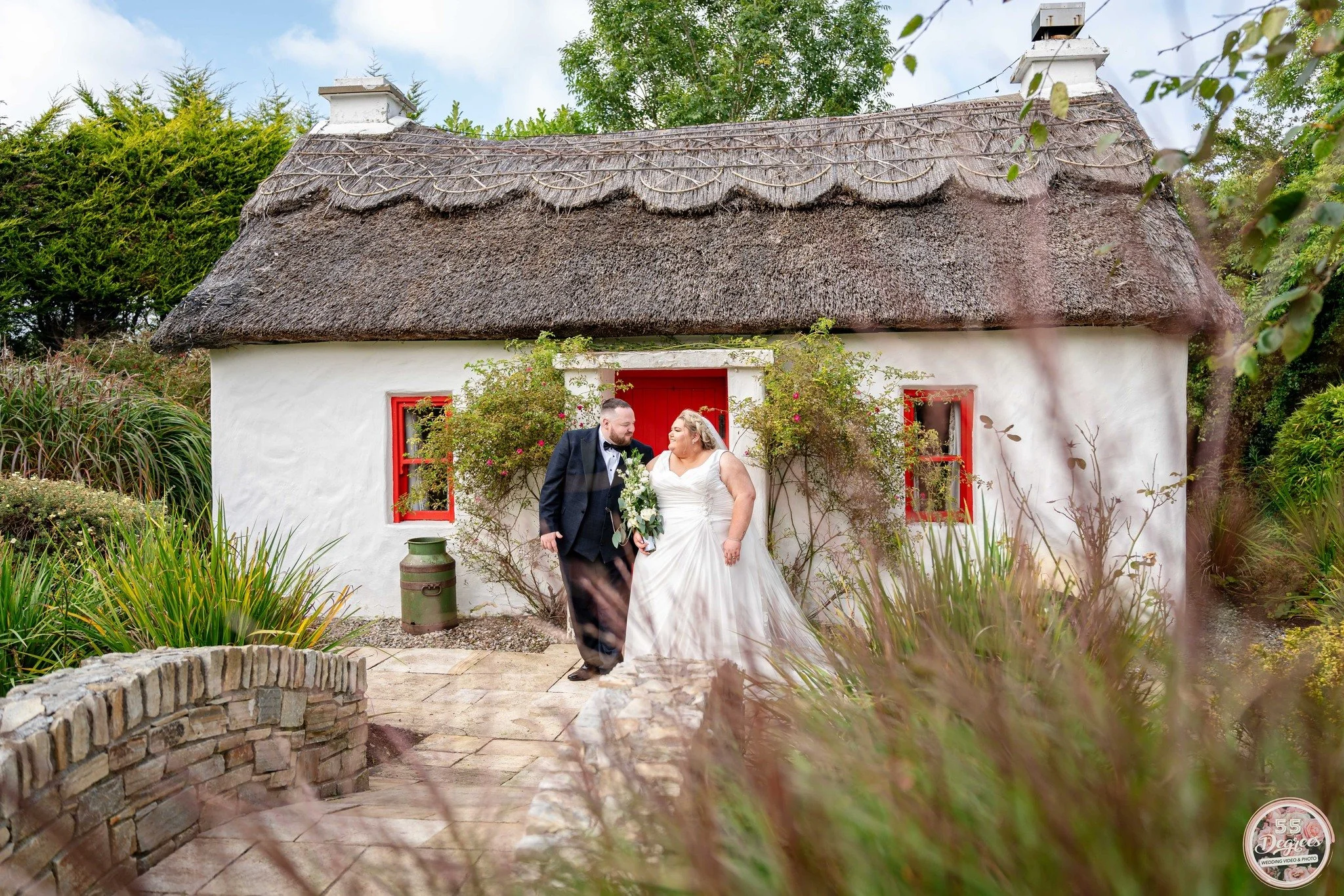 A newlywed couple stands in front of a small white cottage with a thatched roof, red window frames, and lush greenery, sharing a happy moment.