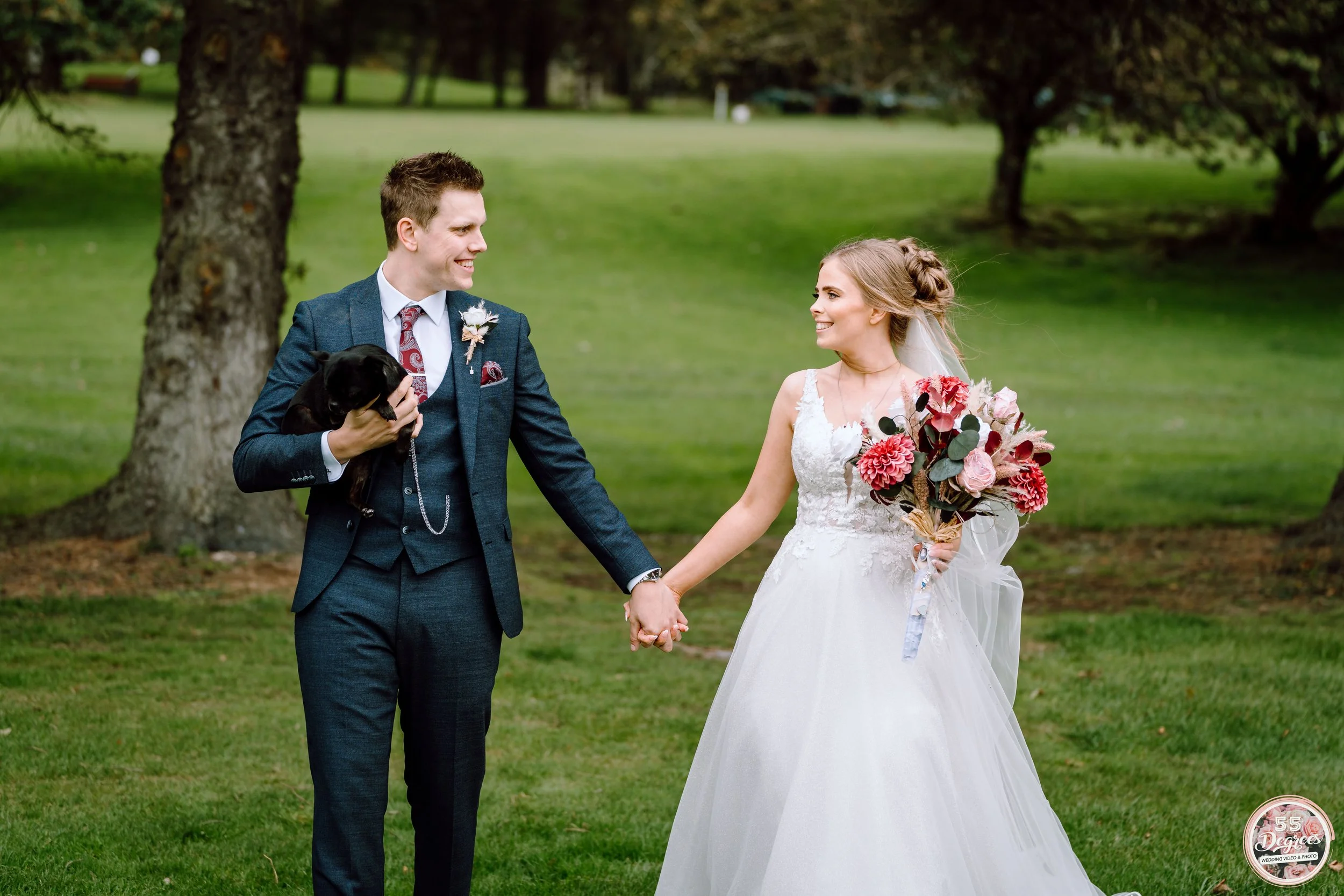 A bride and groom holding hands outdoors on a green lawn, with the groom holding a small black dog, smiling at each other. The bride is wearing a white wedding dress and holding a bouquet of pink and red flowers. There are trees in the background.