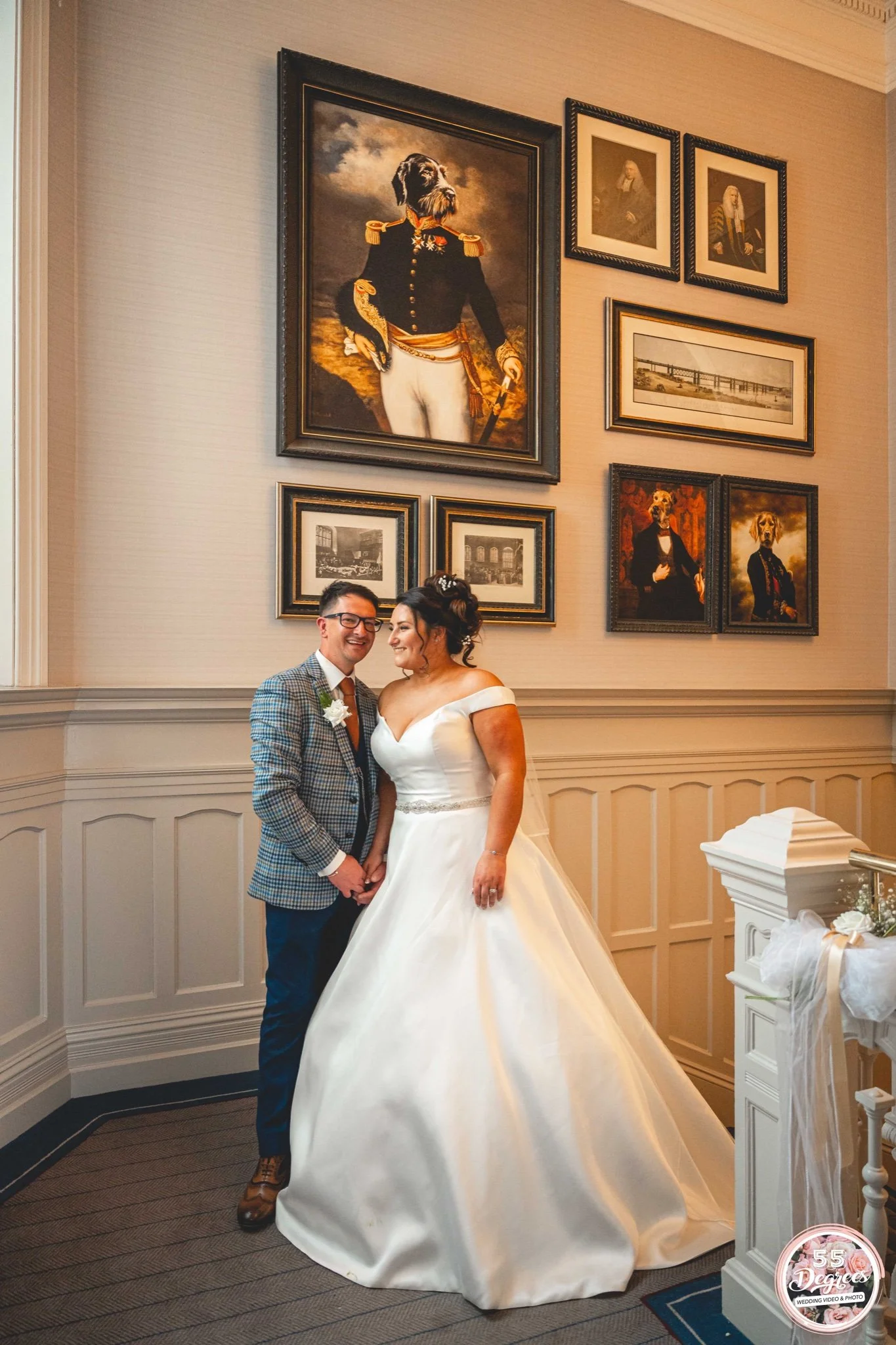 A bride and groom holding hands and smiling at each other indoors, with ornate framed paintings and photographs of historical figures and architecture on the wall behind them.