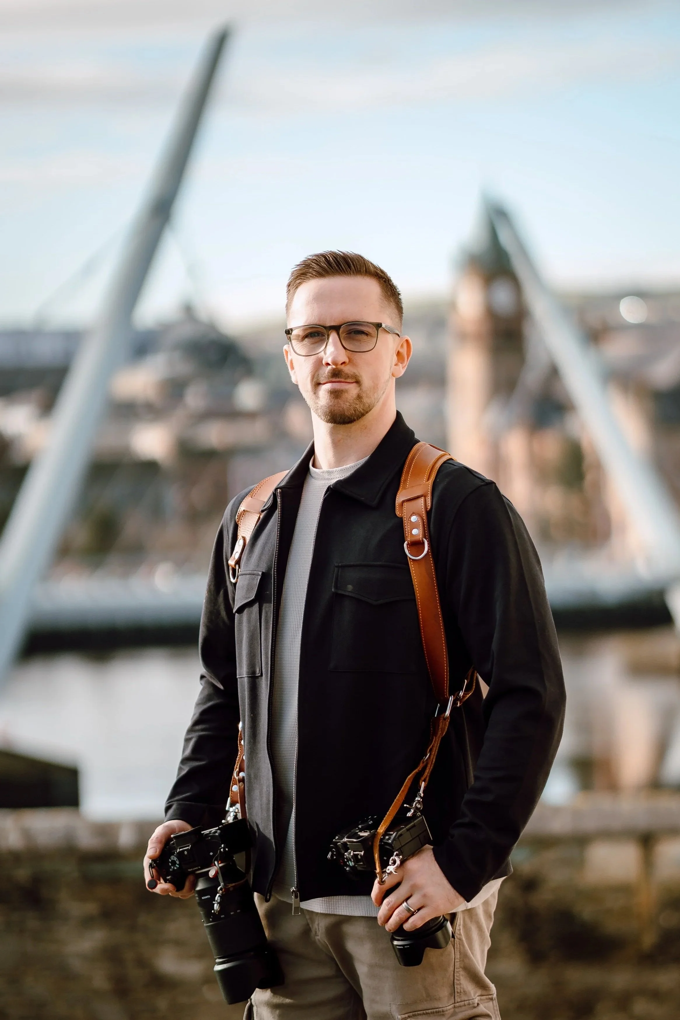 Young man with glasses holding two cameras, standing outdoors near a bridge with a blurred cityscape in the background.