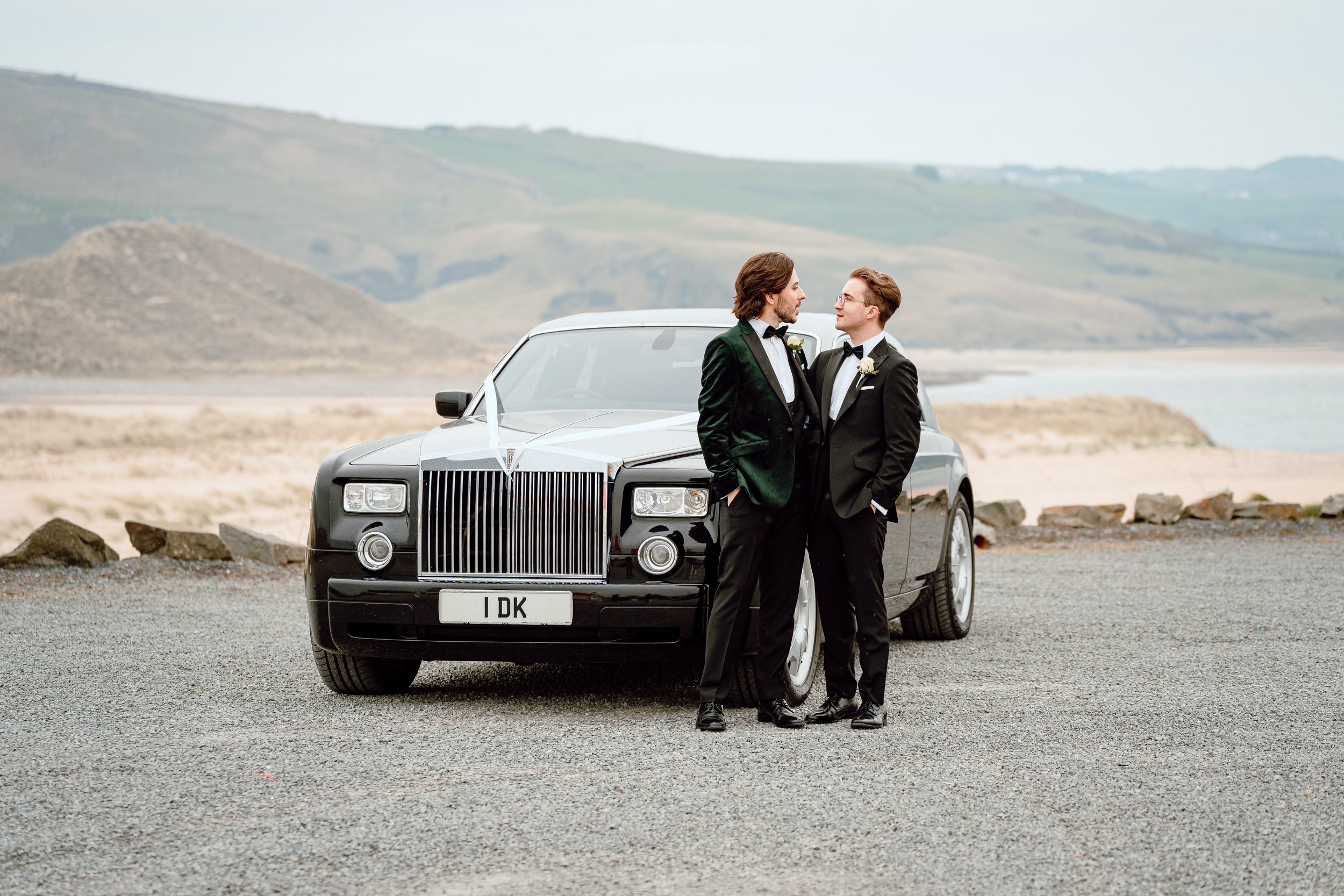 Two men in tuxedos standing close to each other in front of a black luxury car, with a scenic coastal landscape in the background.
