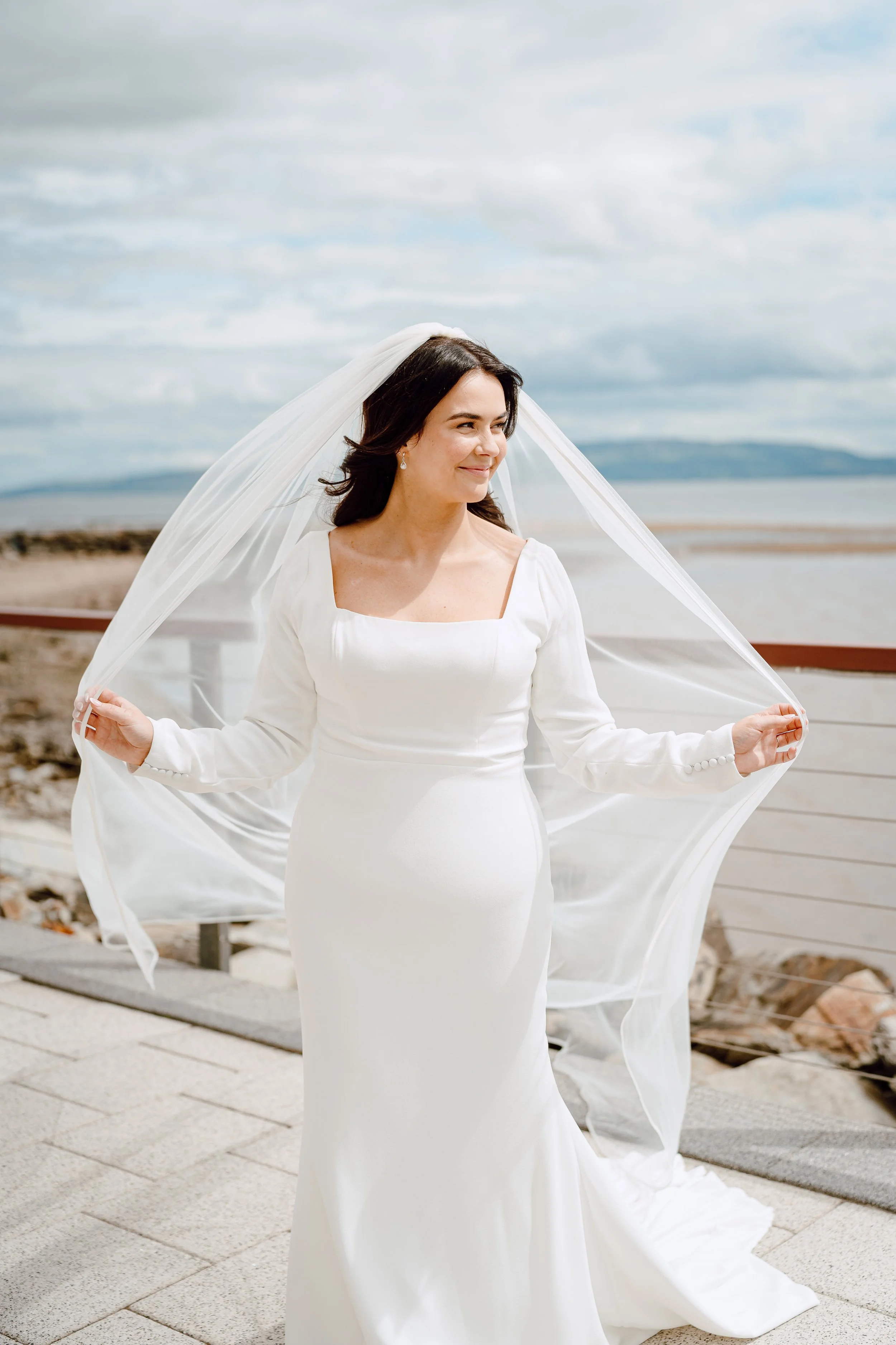 A bride in a white wedding dress standing outdoors near the water, holding her veil, with a cloudy sky in the background.