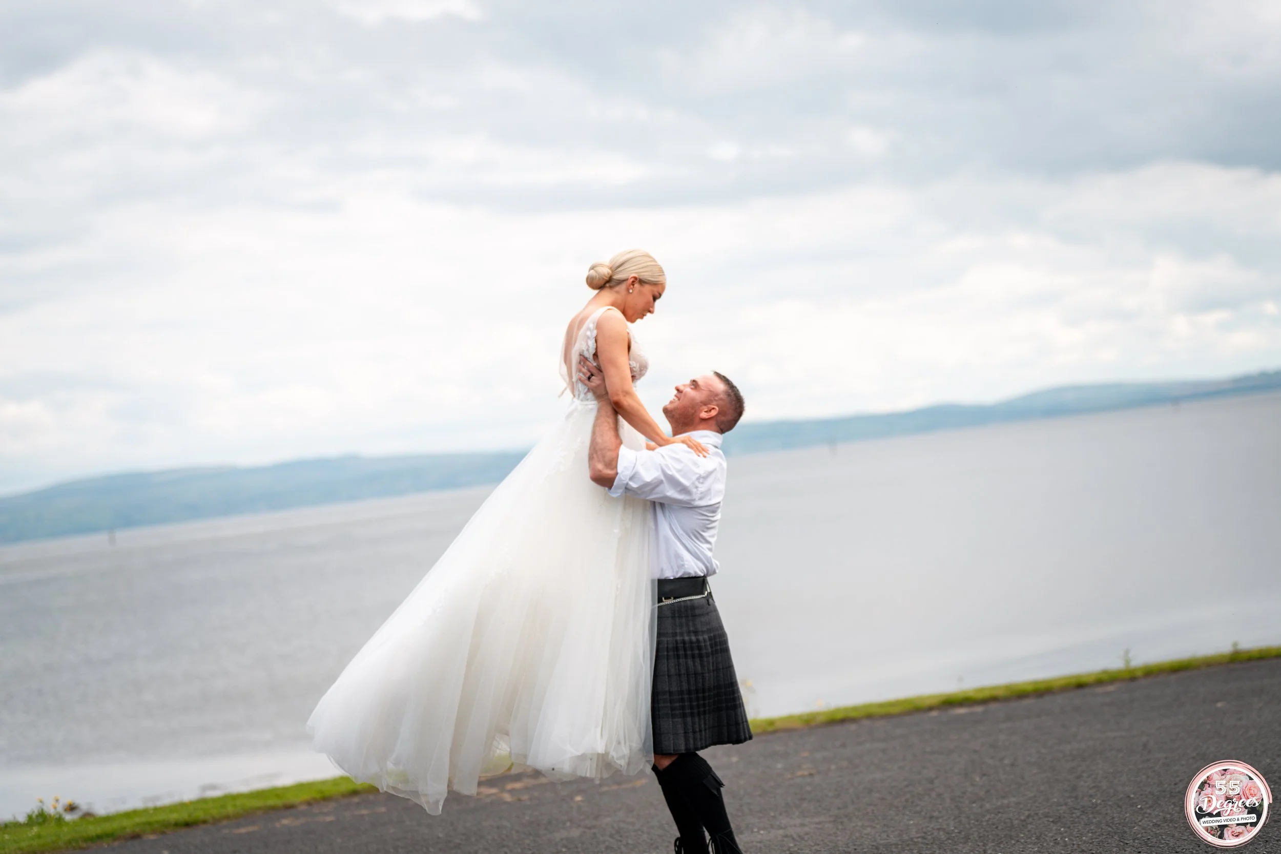 A groom lifting a bride in a white wedding dress outdoors near a body of water, with a cloudy sky overhead.