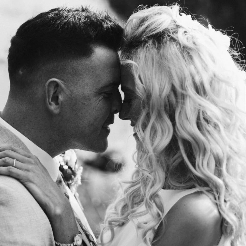 A black and white photo of a couple with their foreheads touching, smiling with their eyes closed. The man has short dark hair, and the woman has long, curly blonde hair. The woman is wearing a ring on her left hand, resting on the man's shoulder.