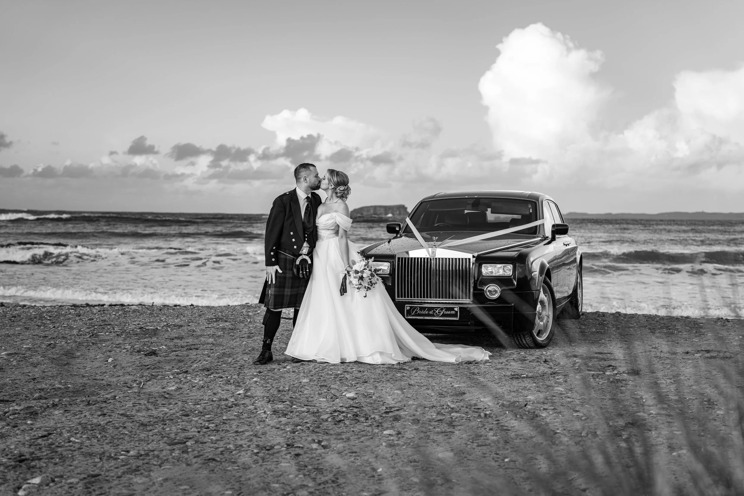 A bride and groom standing on a beach, sharing a kiss in front of a decorated luxury car with a sign that reads 'Bride & Groom' on the front license plate in black and white photograph.