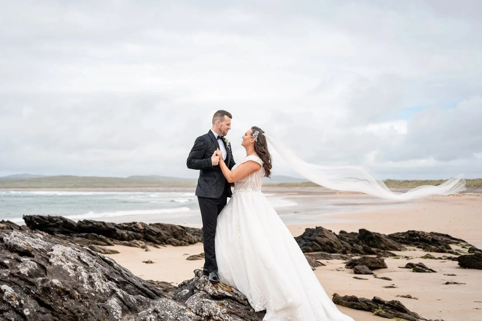 A bride and groom standing on rocks at the beach, looking at each other, with the bride's veil flowing in the wind and the ocean in the background.