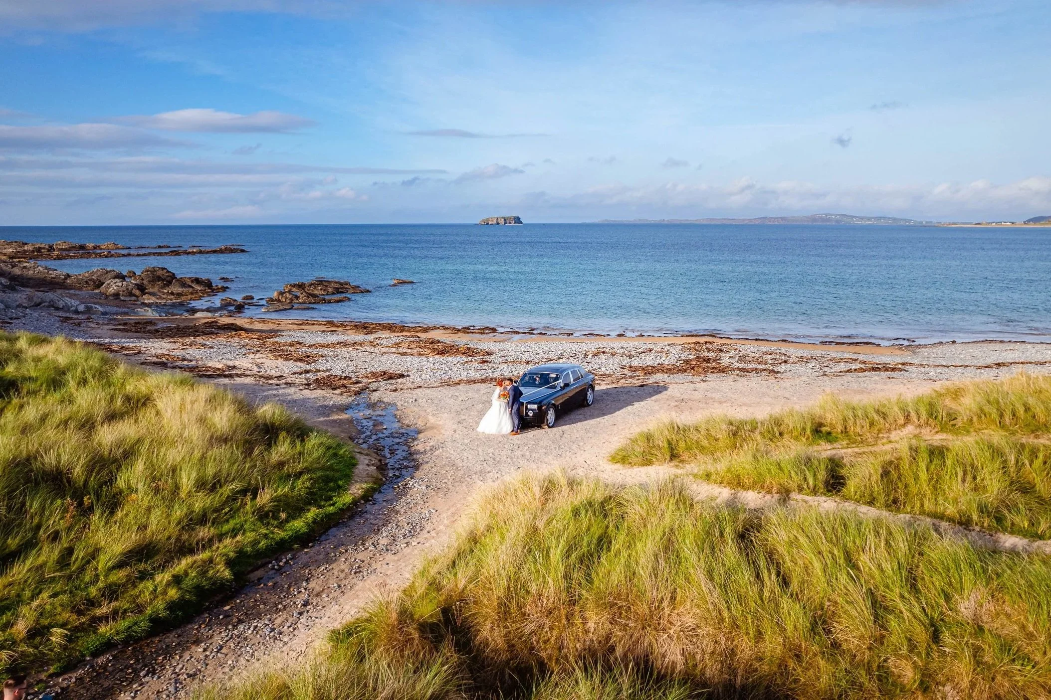 A couple in wedding attire standing next to a black car on a sandy beach with grassy dunes, ocean, and distant islands under a partly cloudy sky.