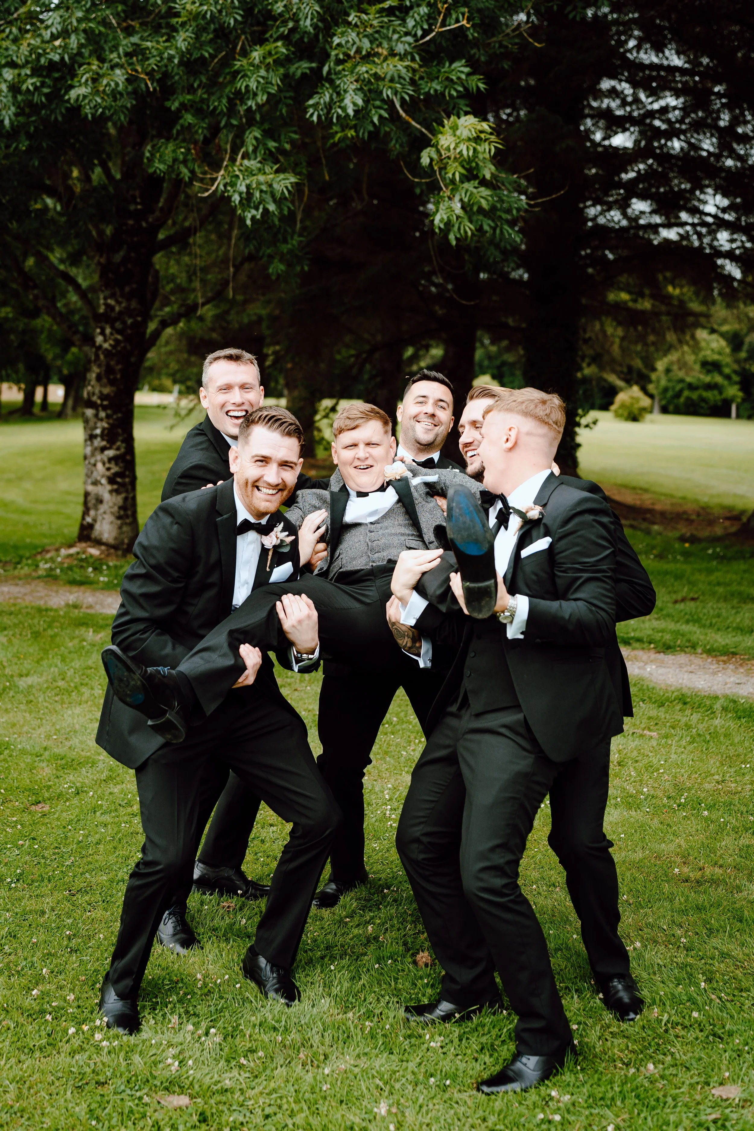 A group of men in tuxedos lifting a man in a suit, smiling and laughing outdoors with trees and grass in the background.