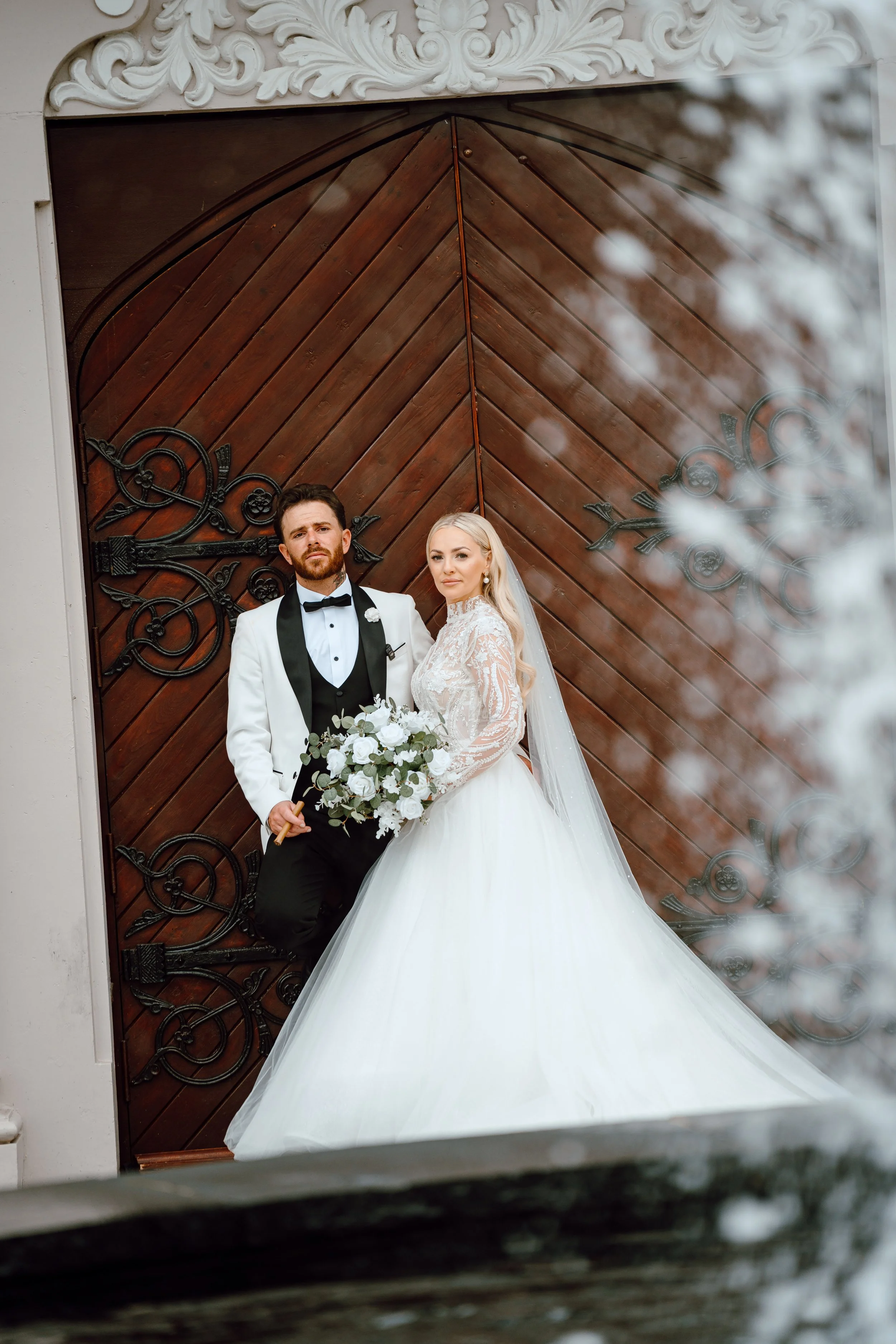 Bride and groom in wedding attire standing in front of a large wooden door with ornate black metalwork, outside the building.
