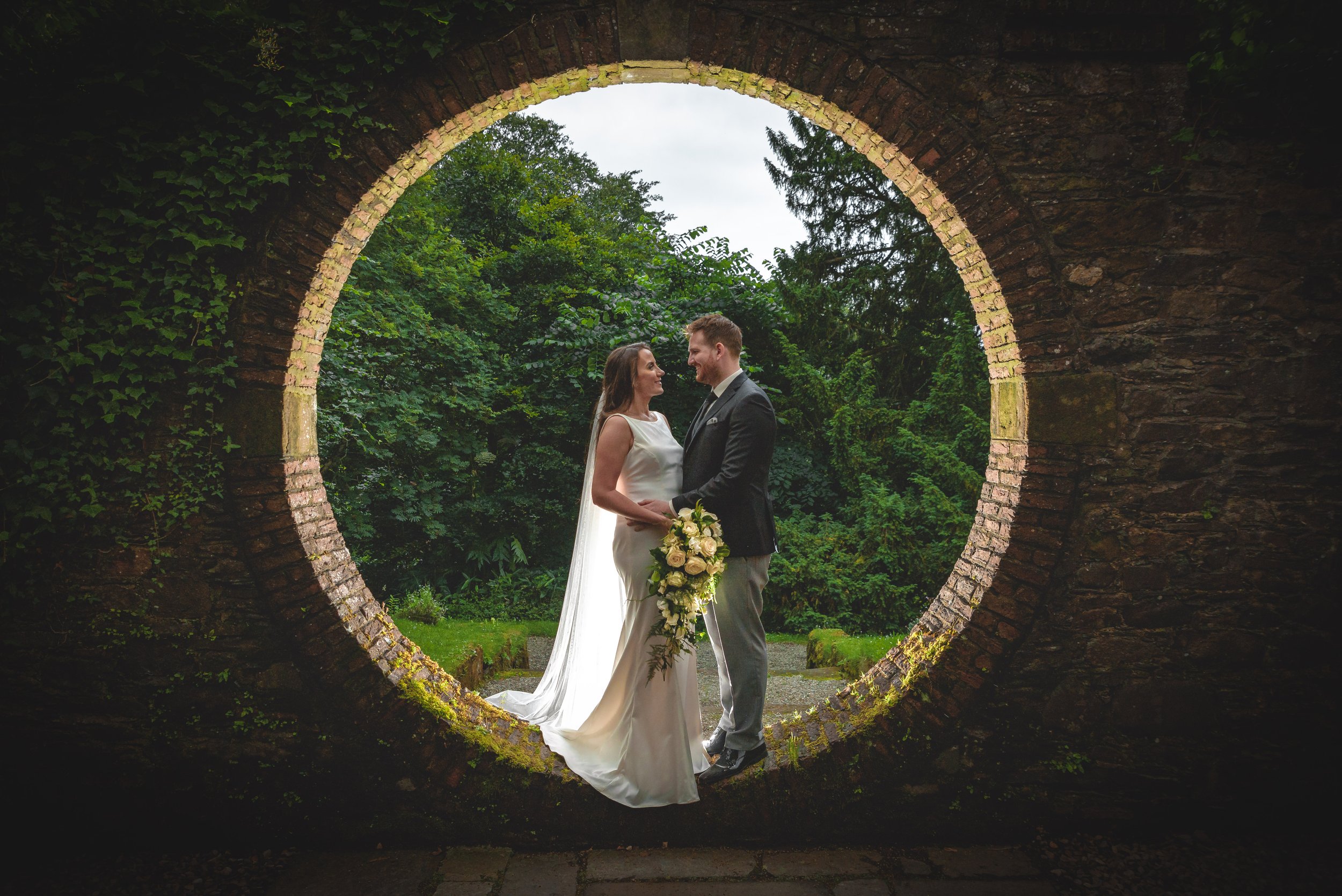 A bride and groom in wedding attire looking at each other, holding hands, and standing inside a circular opening in a brick wall with greenery in the background.