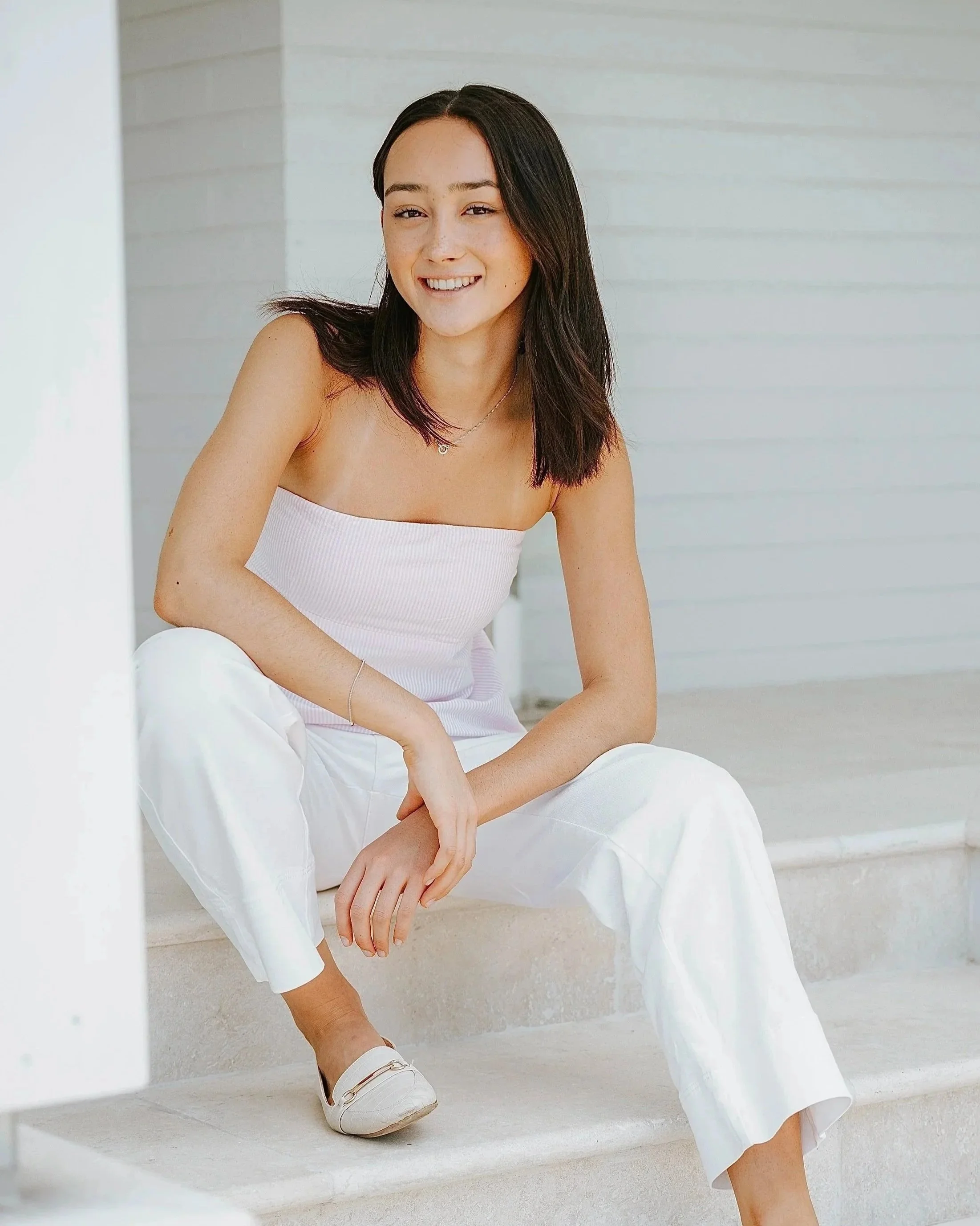 A young woman with dark hair, wearing a pink strapless top and white pants, sitting on stairs outside, smiling at the camera.