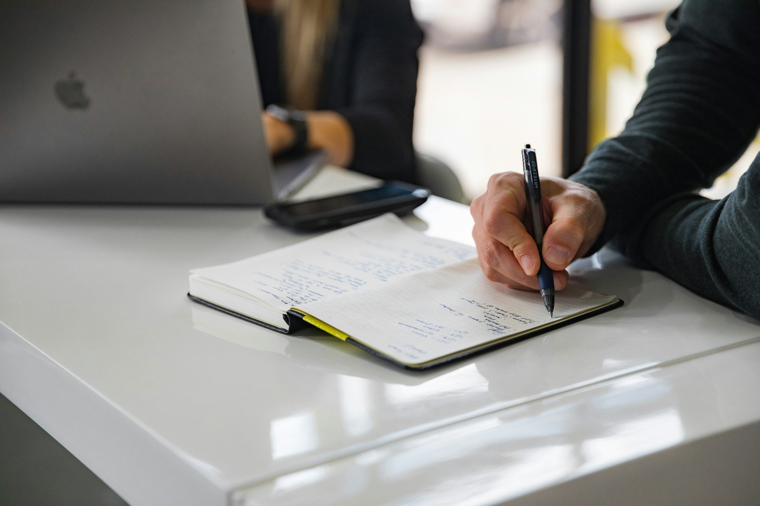 Person writing notes in a notebook on a white desk, with a laptop, smartphone, and another person working in the background.
