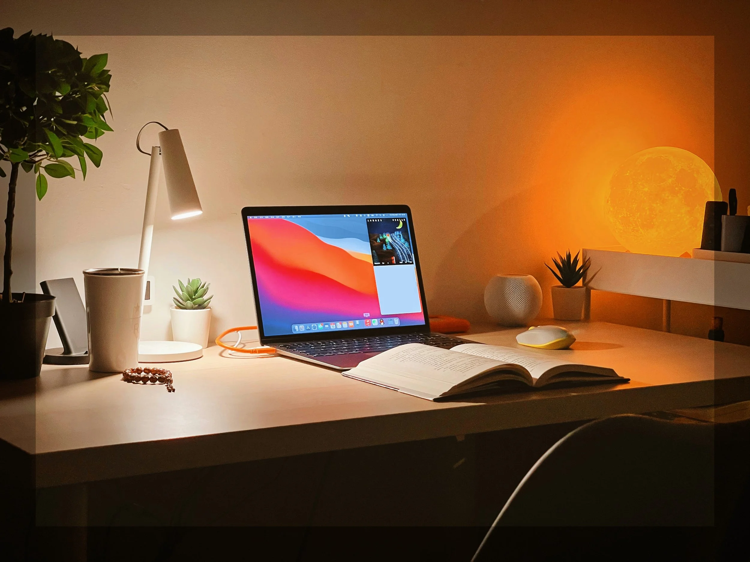 A well-organized desk illuminated by a desk lamp, with a laptop displaying a colorful desktop, an open book, a computer mouse, several small potted plants, a ceramic cup, a beaded bracelet, and a glowing full moon projection on the wall