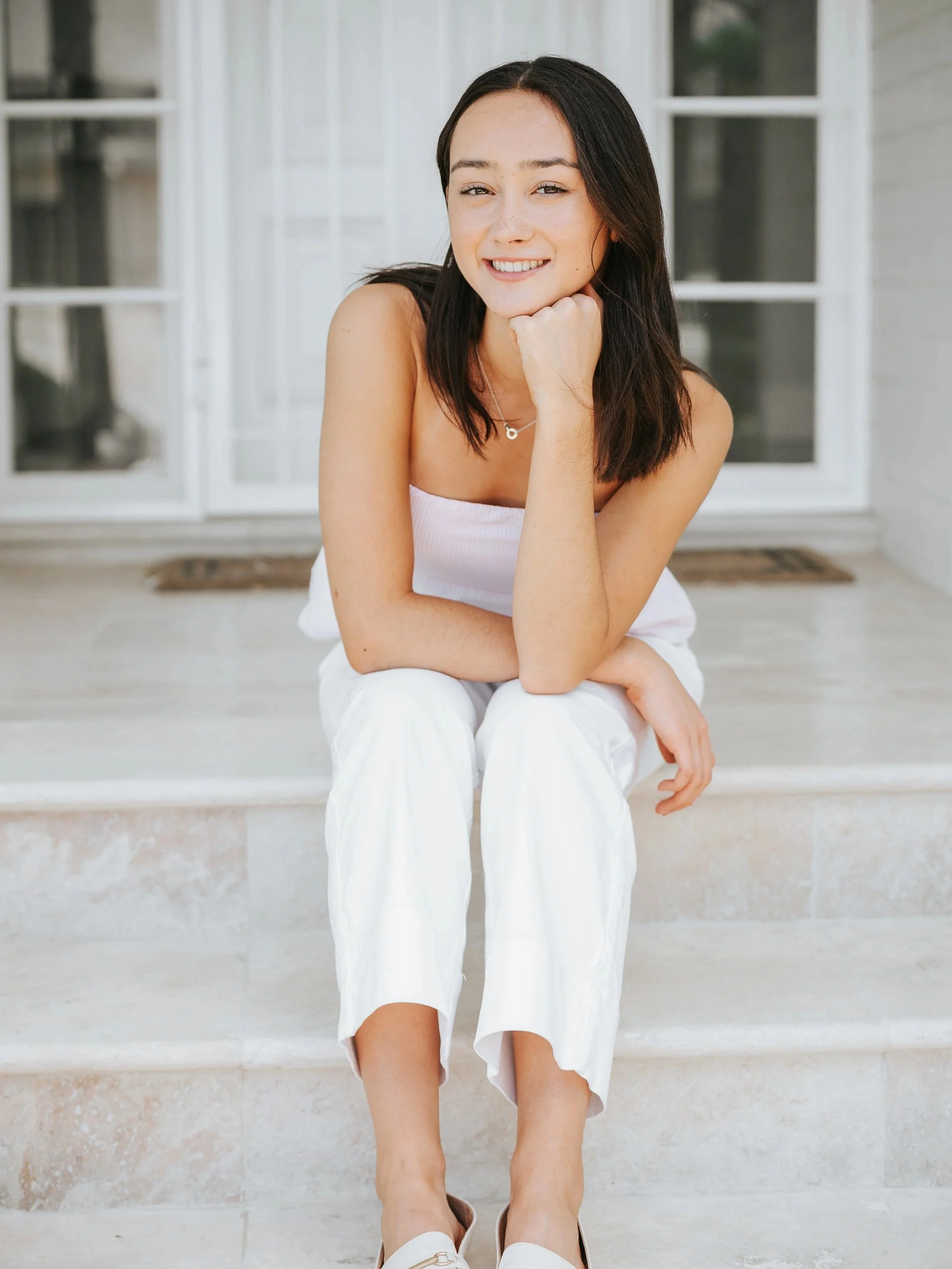 A young woman with dark hair, wearing a white strapless top, white pants, and light-colored shoes, sits on the steps of a porch, smiling at the camera with her chin resting on her hand.