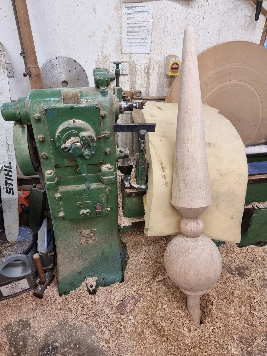 A woodworking lathe machine with a partially turned wooden spindle attached, surrounded by sawdust in a workshop.