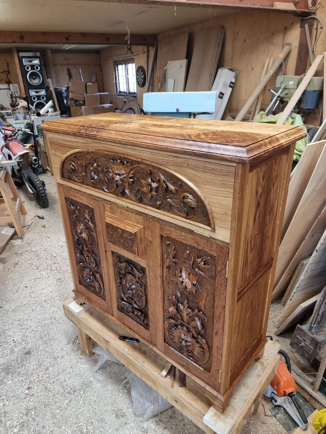 A handcrafted wooden cabinet with ornate carved floral designs on the front in a woodworking shop.