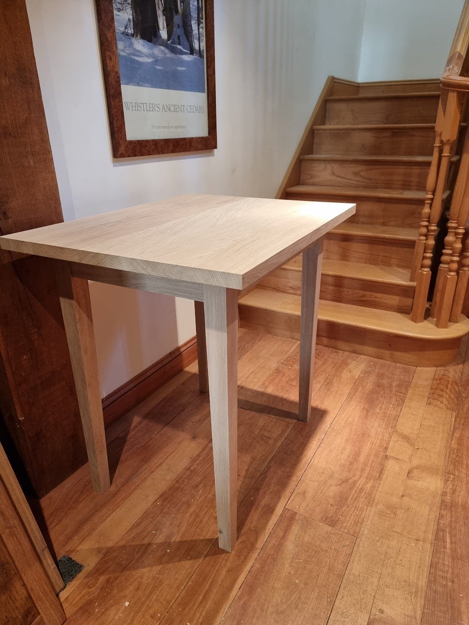 A small wooden table in a corner with stairs in the background and a framed poster on the wall.