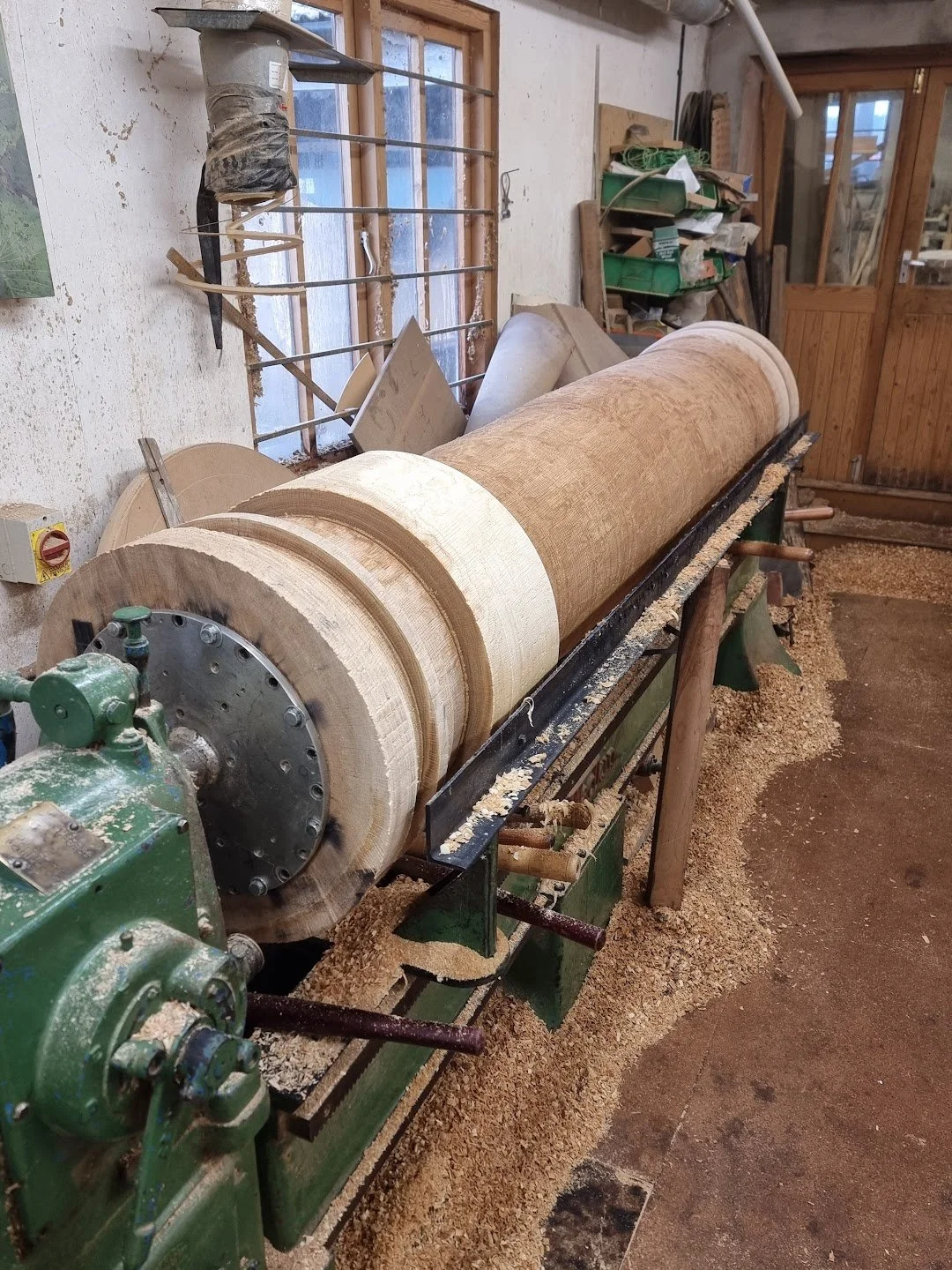 A woodworking lathe machine with a piece of wood mounted, surrounded by wood shavings in a workshop.