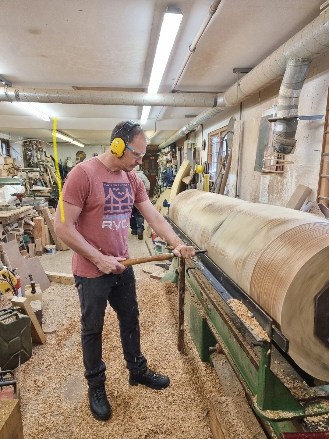A woodworker in a workshop is shaping a large piece of wood on a lathe, wearing safety glasses and hearing protection.