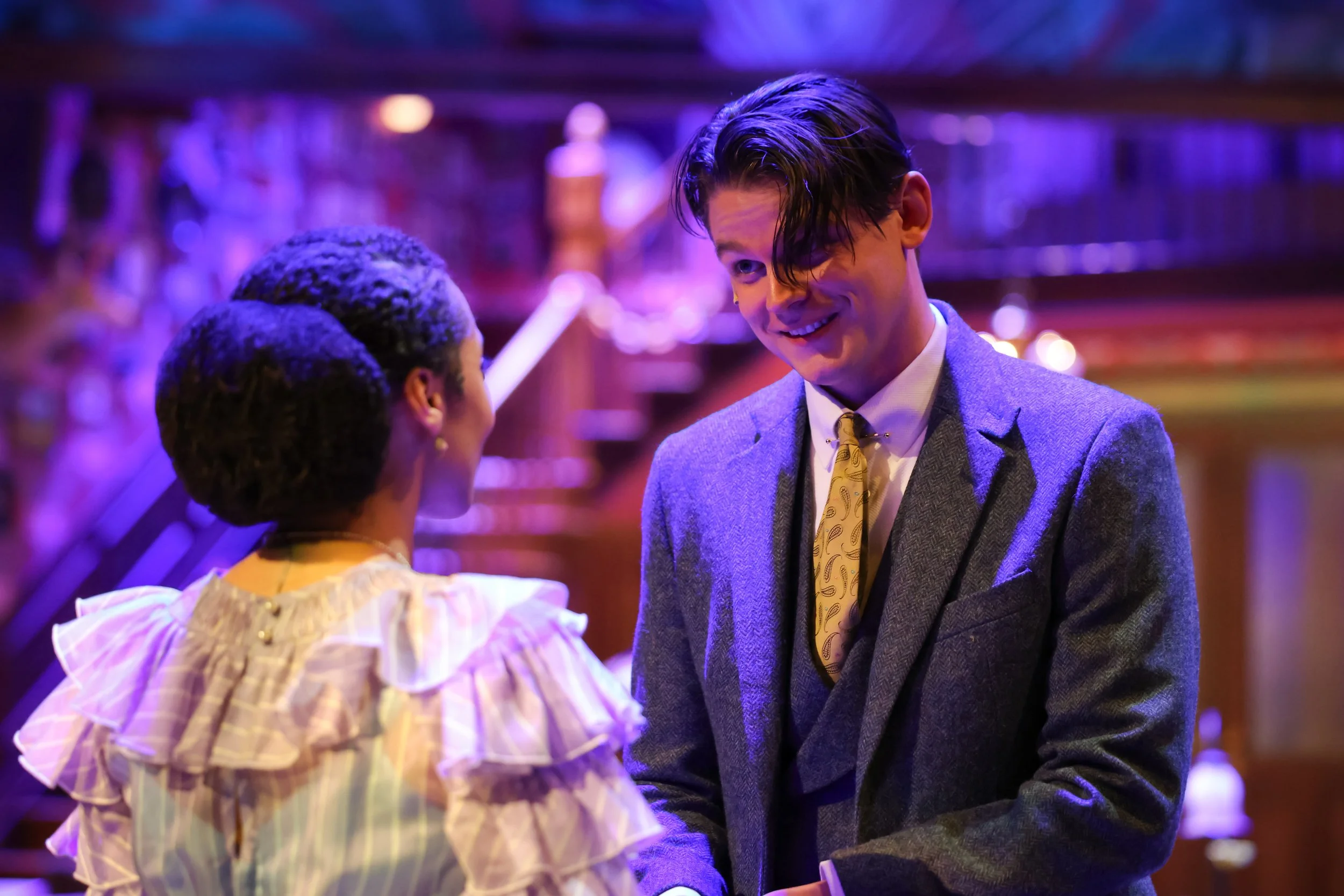 Reez Bailey, actor, performing in You Can't Take It With You at PlayMakers Repertory Company. A man with dark, wavy hair smiling and talking to a woman in a fancy dress in a warmly lit room with purple lighting.