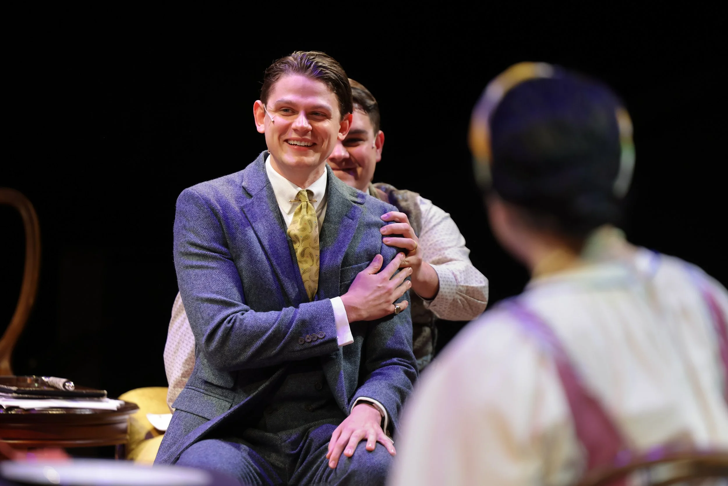 Three men on stage, two are visible in the foreground, one is in the background, with the person in the foreground, right, wearing a dark patterned hat and patterned shirt. The man in the center is sitting, wearing a blue suit jacket, white shirt, and a yellow tie, smiling and holding his hand on his chest, while the man behind him has a hand on his shoulder.