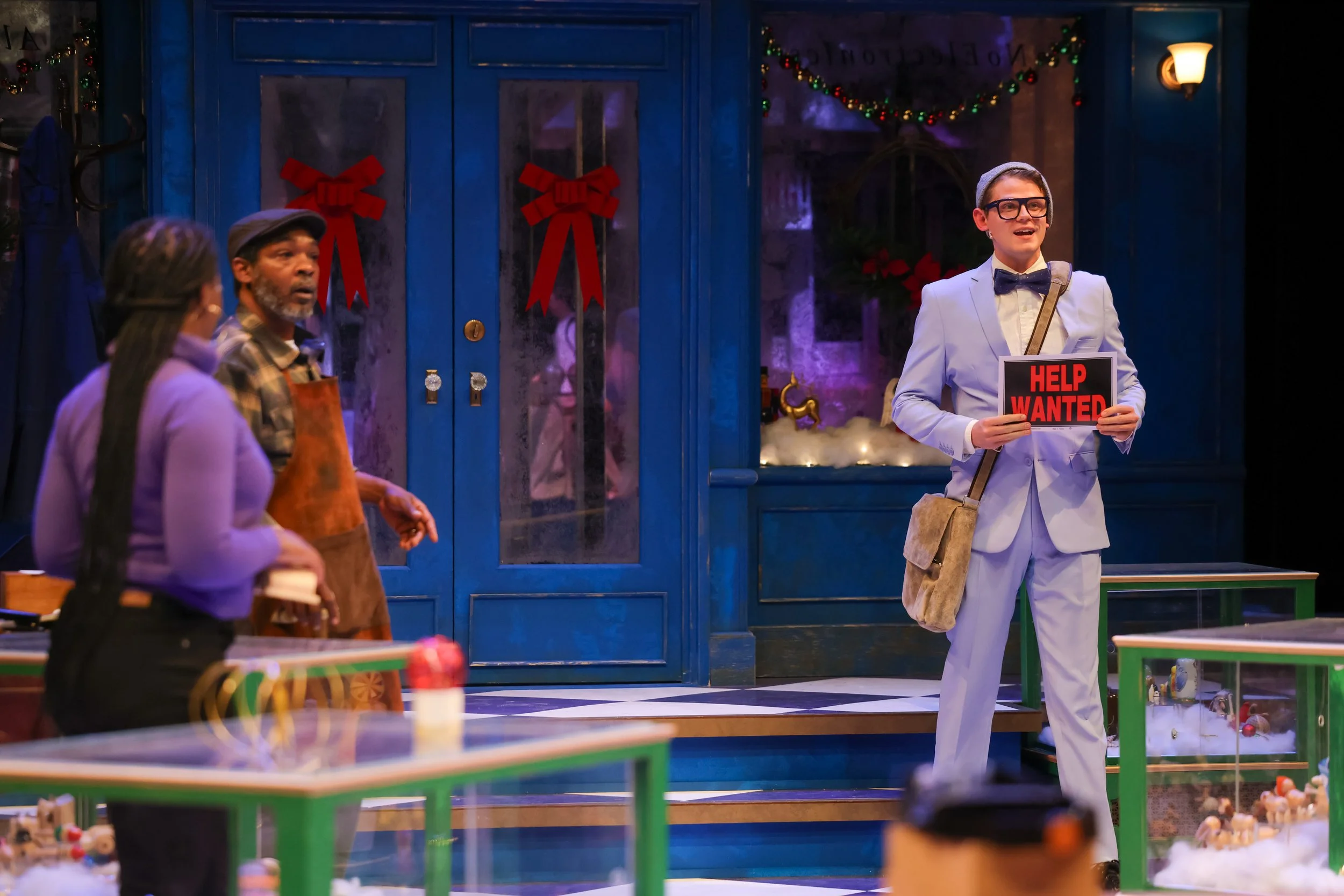 A man in a light blue suit holding a 'Help Wanted' sign stands on a stage decorated with Christmas ornaments. Two people, a woman and a man, are also on stage, facing the man in the suit. The background features a blue door with red bows and Christmas garlands, and the foreground shows glass display cases with toys.