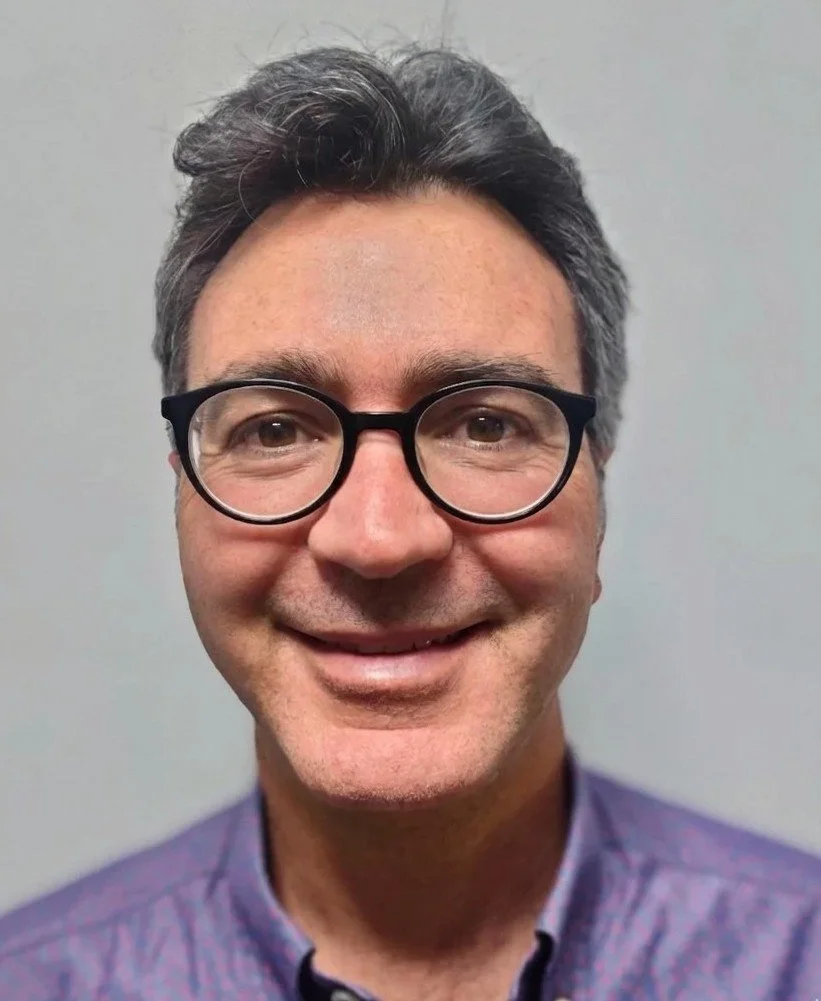A middle-aged man with dark hair, glasses, and a light purple shirt smiling against a plain gray background.