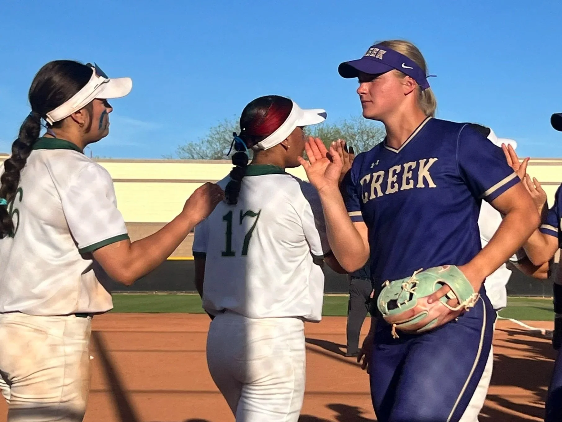Softball players from opposing teams exchange handshakes after a game, showing sportsmanship on the infield under clear skies.
