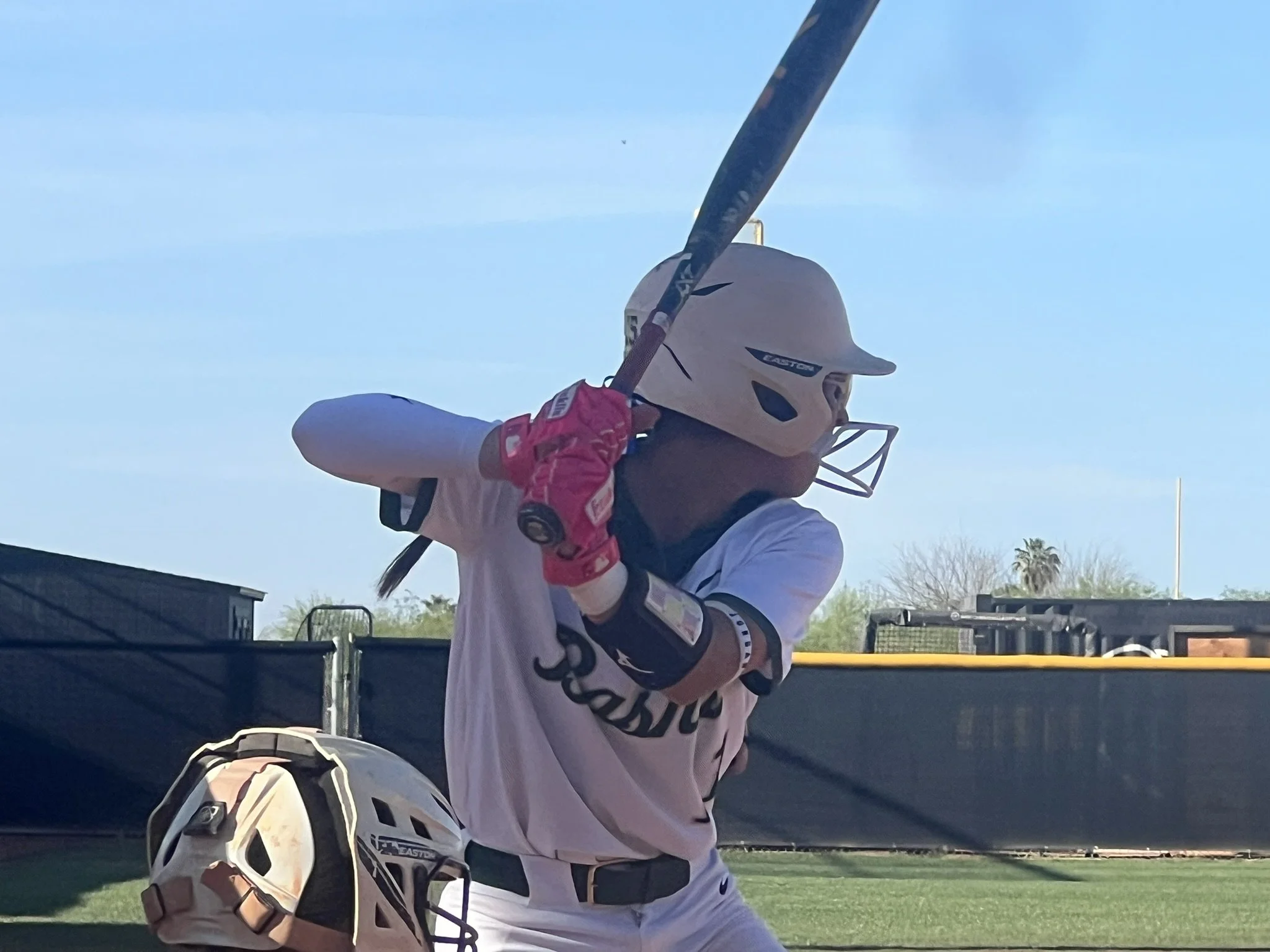 A softball player in a white uniform swings a bat during batting practice, wearing a helmet and pink batting gloves.