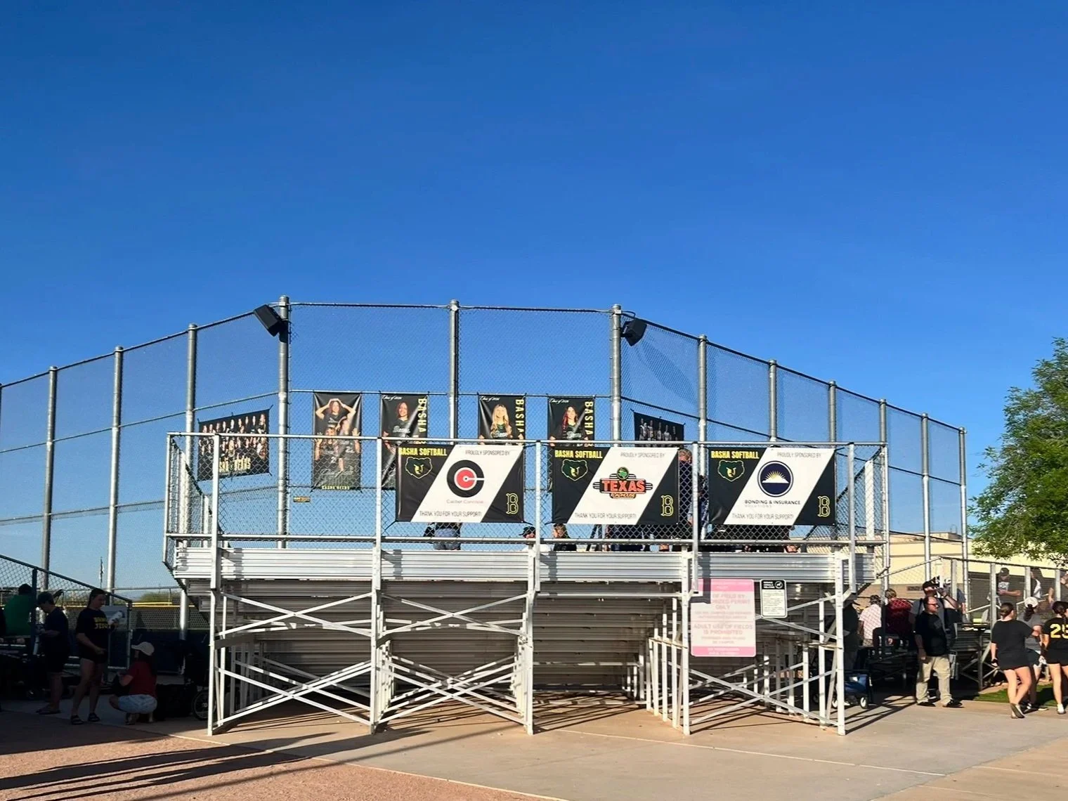 Fans gather around metal bleachers at a high school softball field, with banners and signage displayed along the backstop.