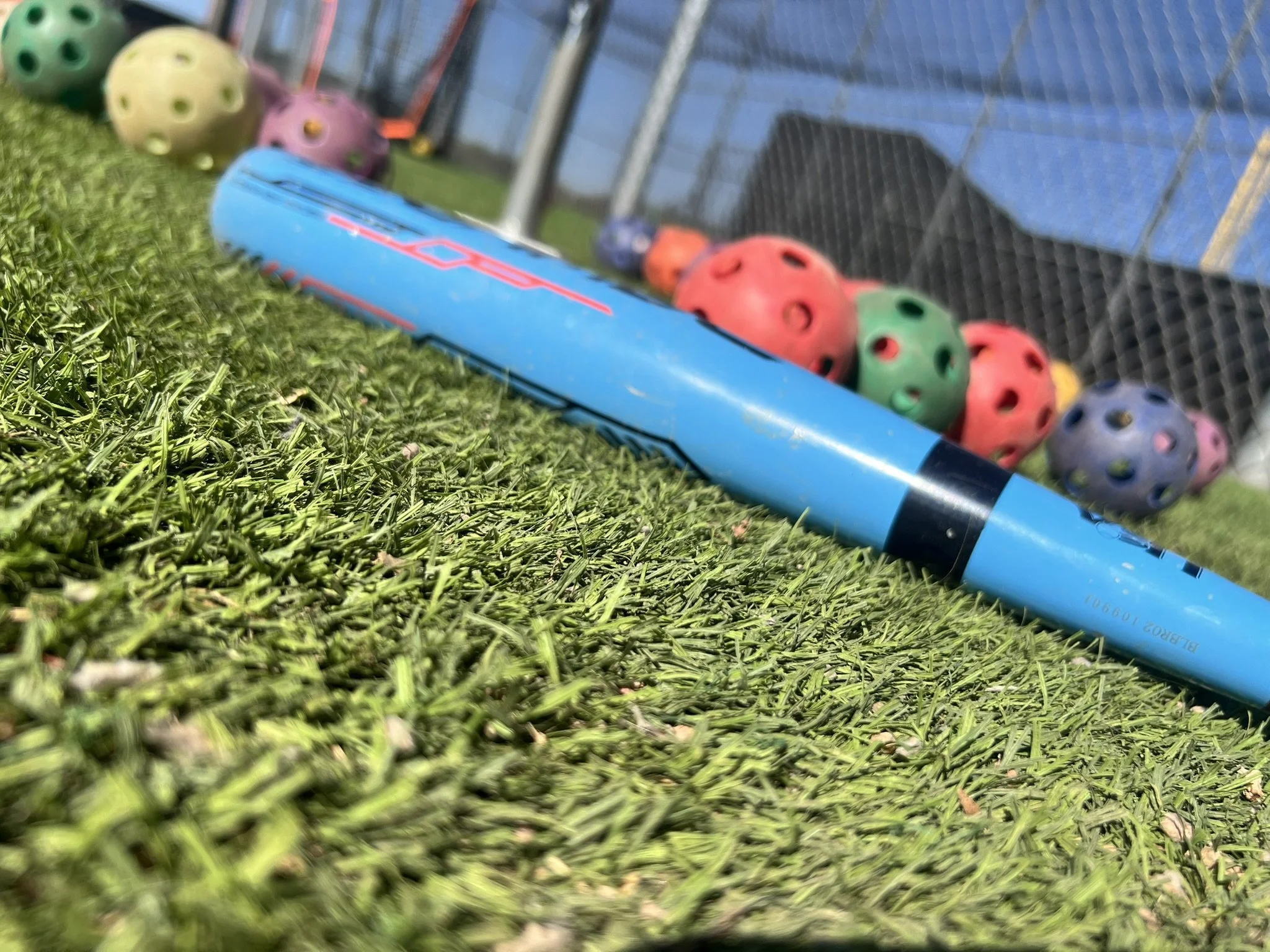 A bat lies on artificial turf beside colorful training balls near a practice net, captured at an angled perspective.