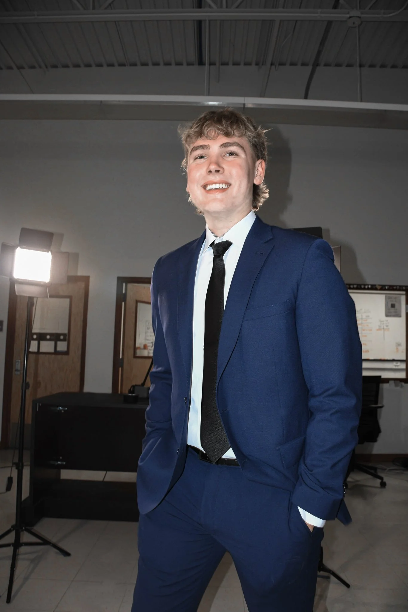 Jayden Riecken in a blue suit, white shirt, and black tie standing indoors near a photography setup, smiling at the camera.