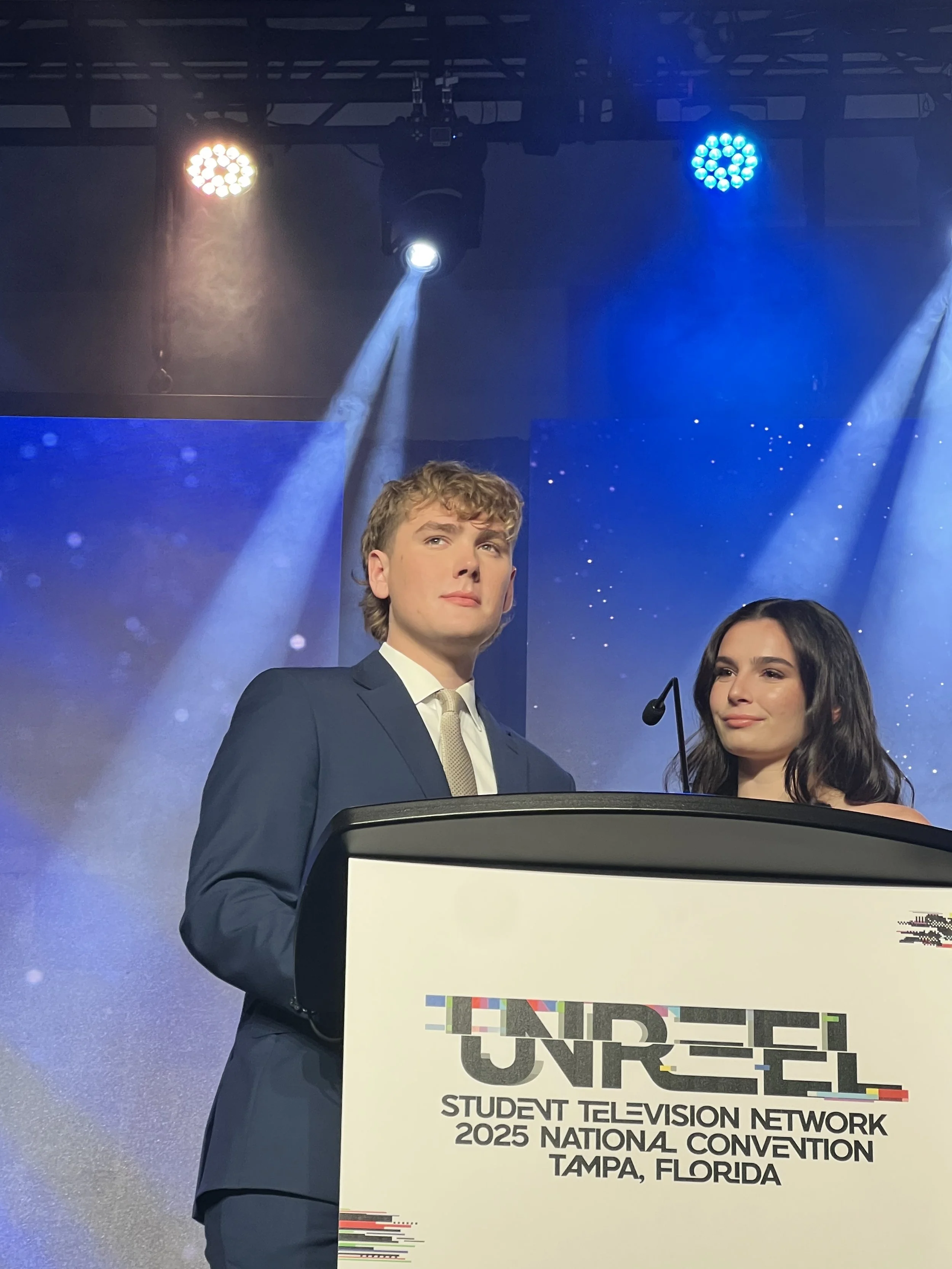 Jayden Riecken and young woman standing behind a podium with the UNREEL logo, during the 2025 National Convention for the Student Television Network in Tampa, Florida, with stage lights shining down.