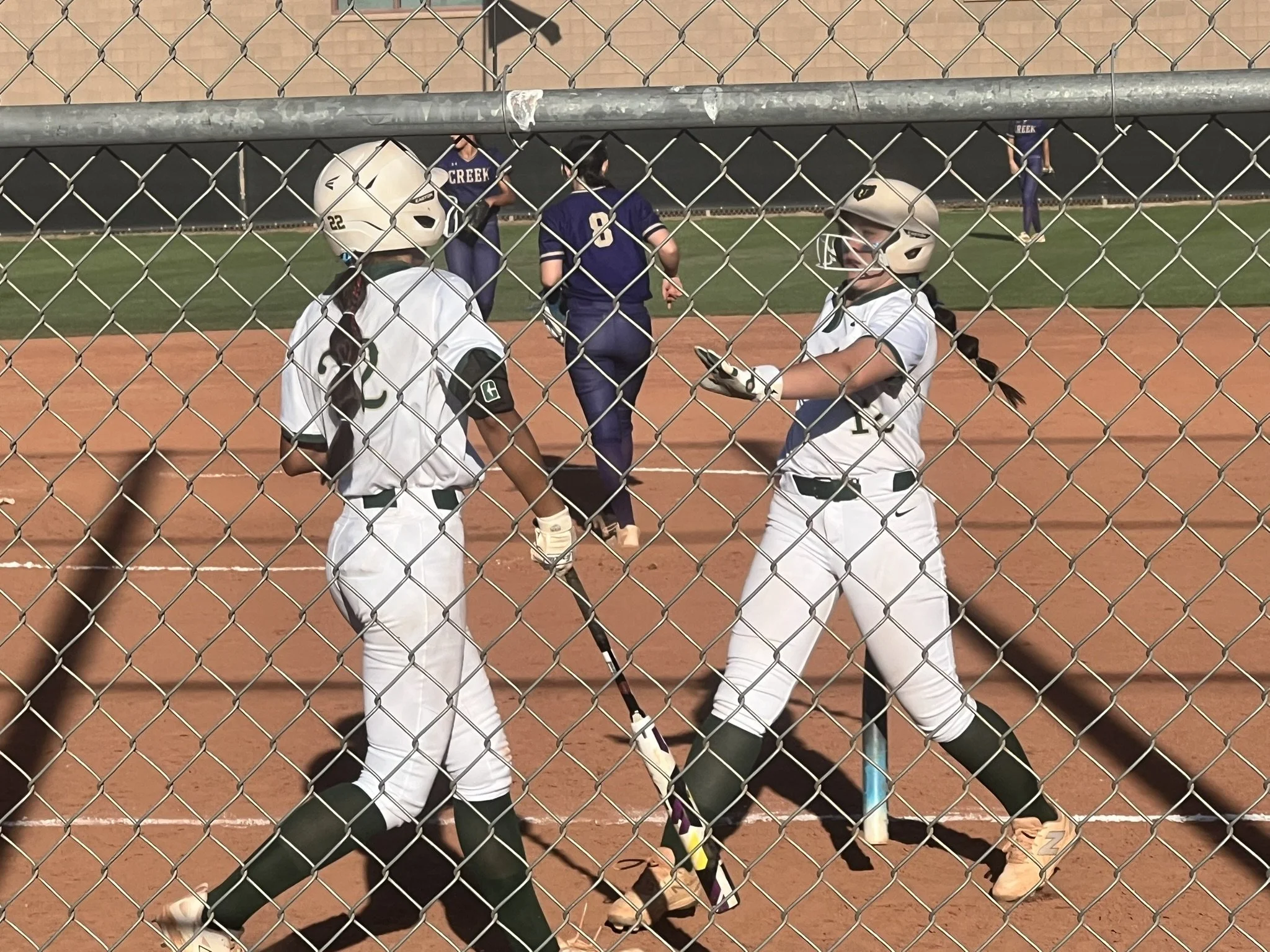 Two softball players in white uniforms exchange a bat near home plate as teammates and opponents stand in the background.