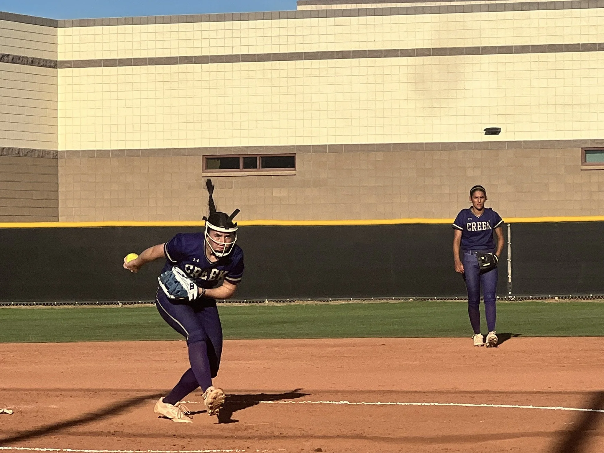 A pitcher in a dark uniform releases the ball during a game, with an infielder positioned behind her on the dirt field.