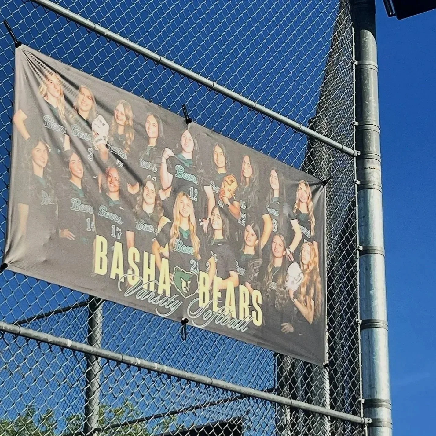 A banner featuring the Basha Bears varsity softball team hangs on a chain-link fence against a clear blue sky.