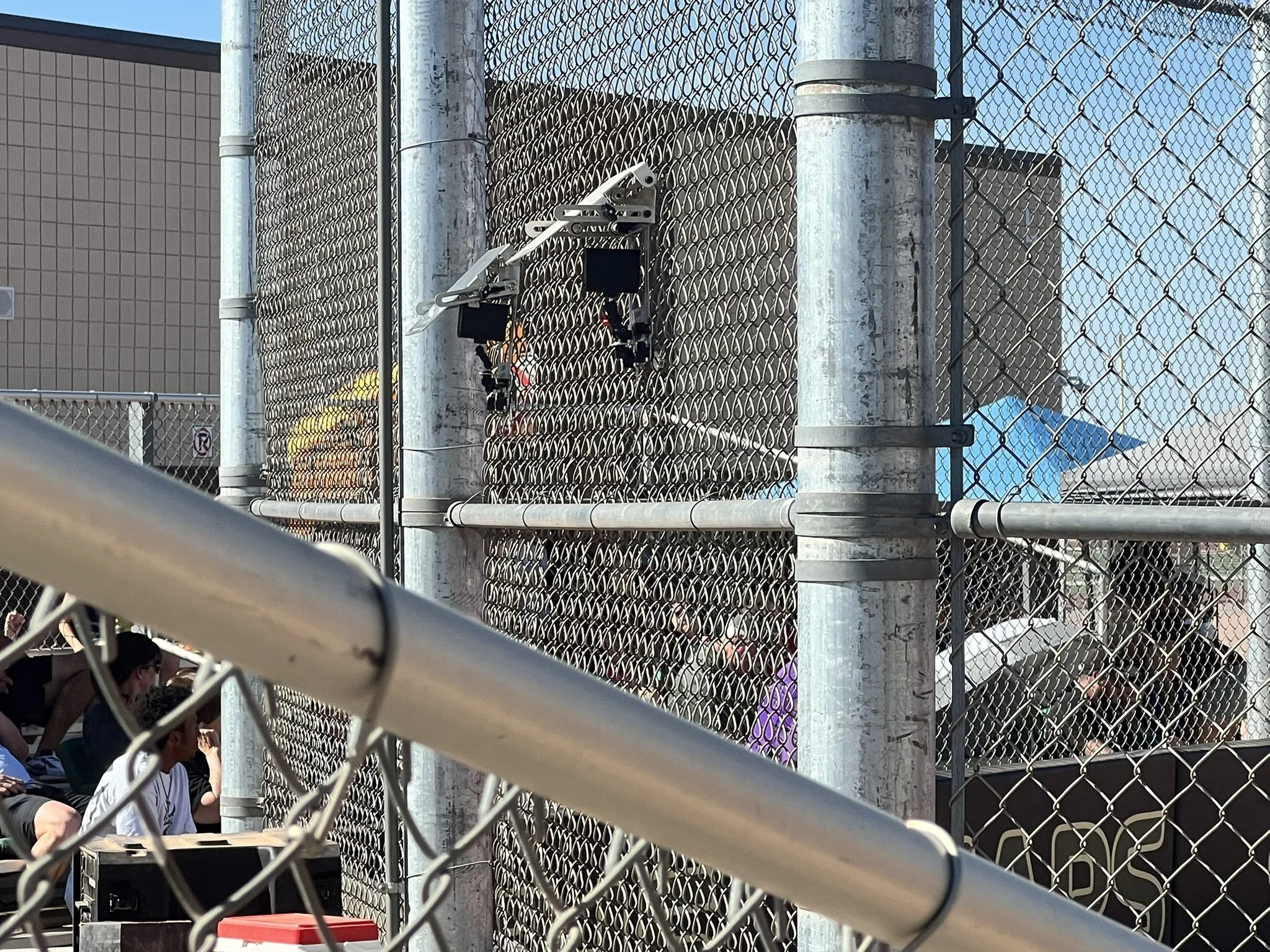 Broadcast cameras mounted on a fence overlook the field, with spectators seated in the background.