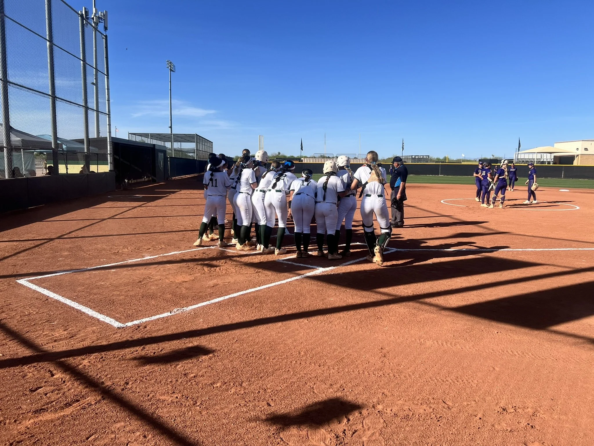 A group of softball players in white uniforms huddle near home plate while the opposing team gathers in the distance.