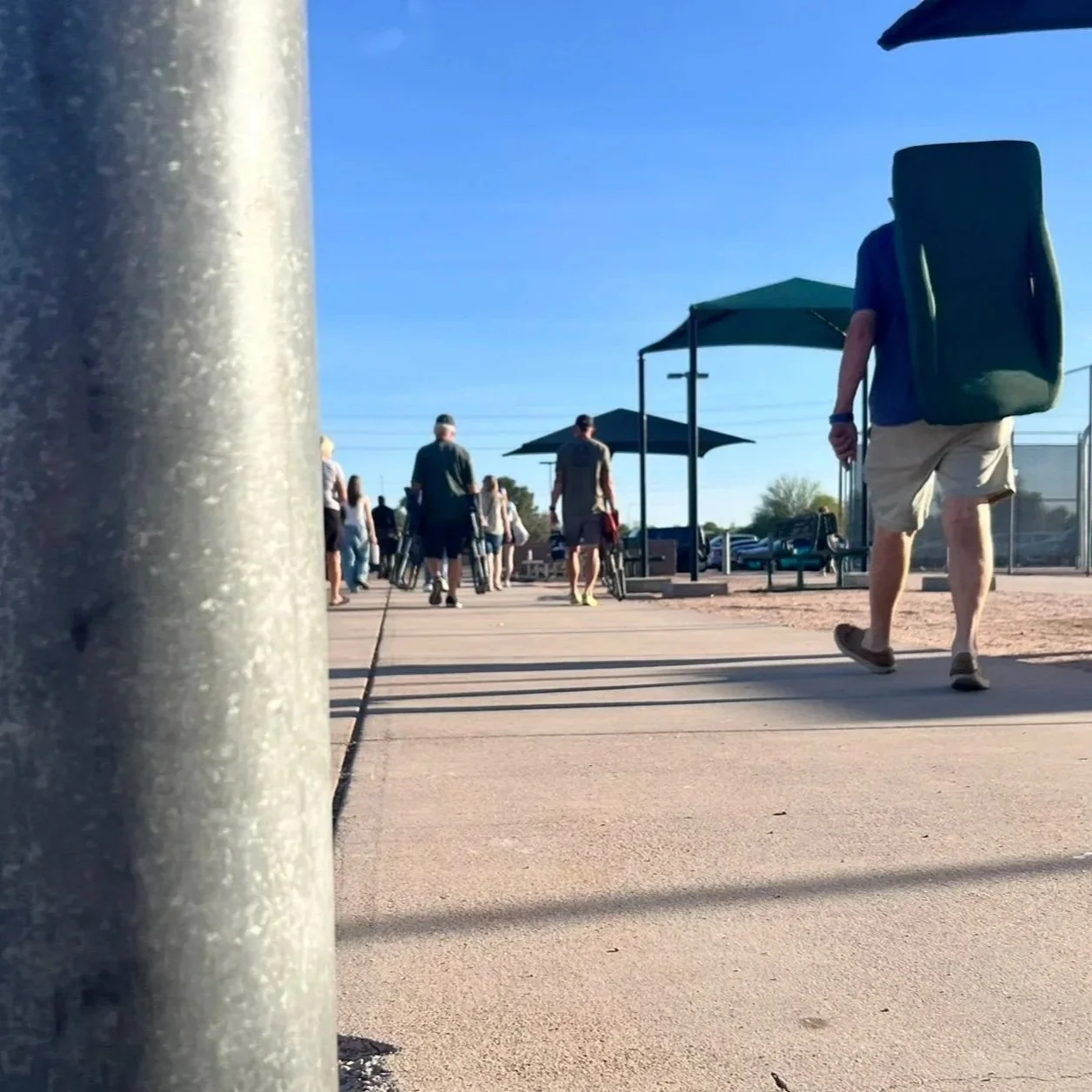 Spectators walk along a concrete path toward the softball complex, with shade structures and fencing lining the field.