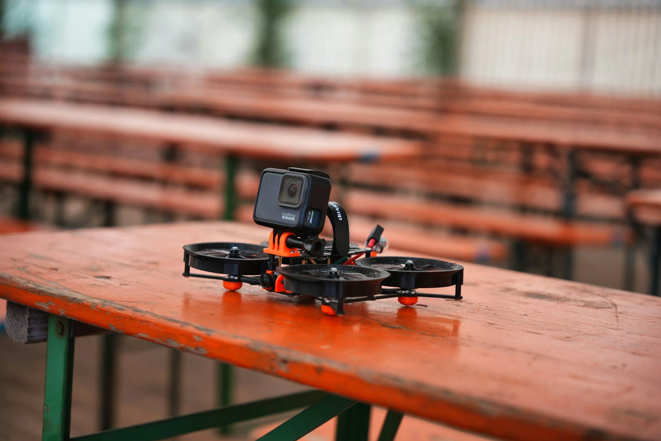 A small drone with a GoPro camera mounted on top is on an orange wooden picnic table with green legs, in an outdoor setting with blurred wooden benches in the background.