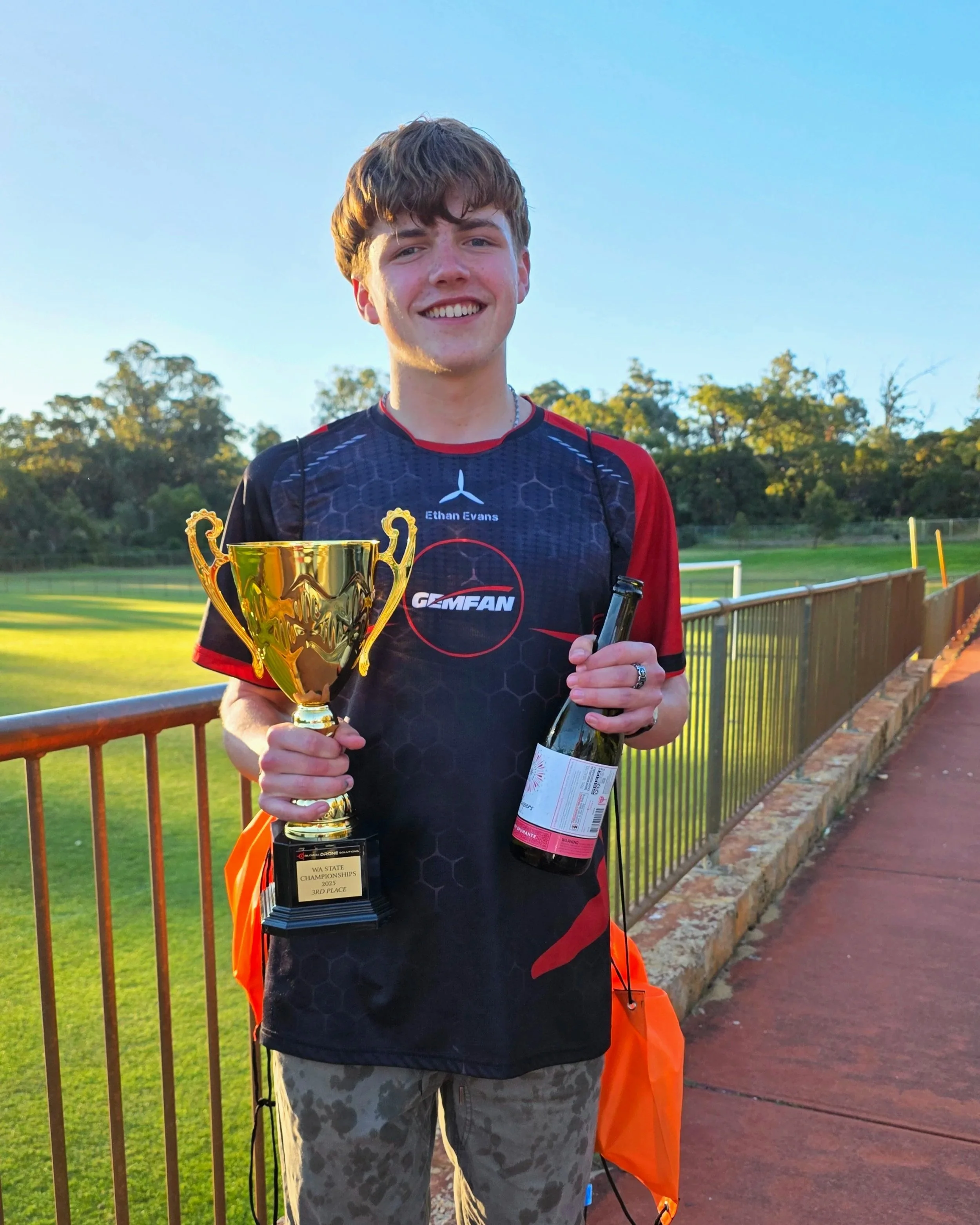 A young man holding a trophy and a wine bottle, smiling outdoors on a sports field with a clear sky.