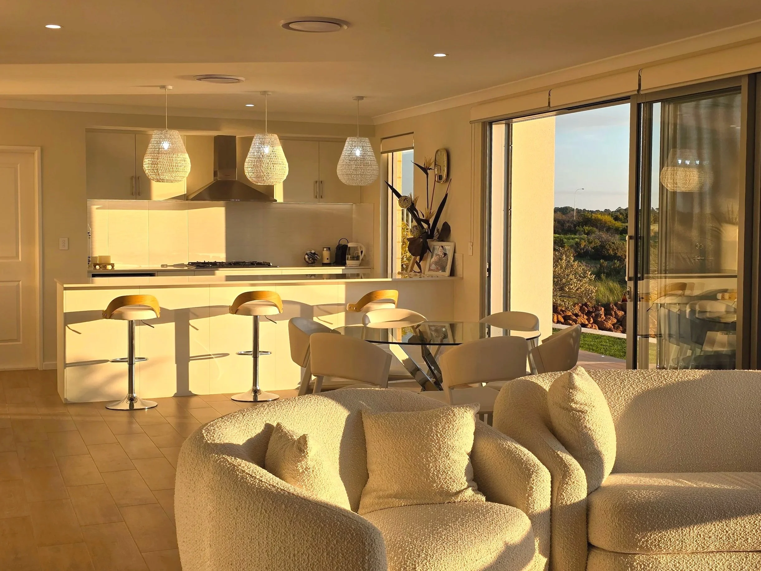 Living room with a white sofa, glass dining table with white chairs, and a kitchen with a white island, three yellow bar stools, and pendant lights, illuminated by warm sunlight through large glass sliding doors showing an outdoor landscape.