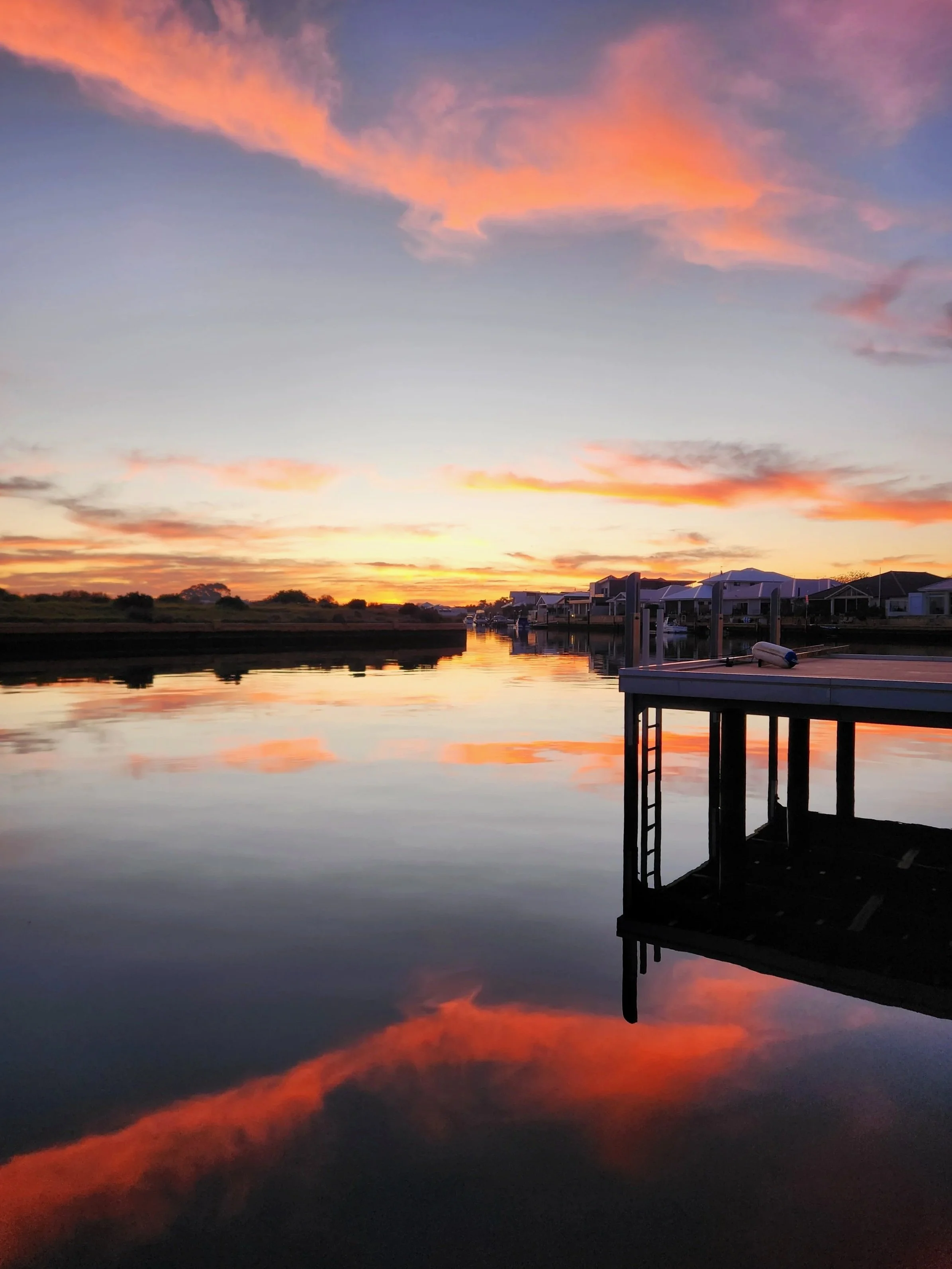 Sunset over calm water reflecting colorful sky, with houses and a dock in the foreground.